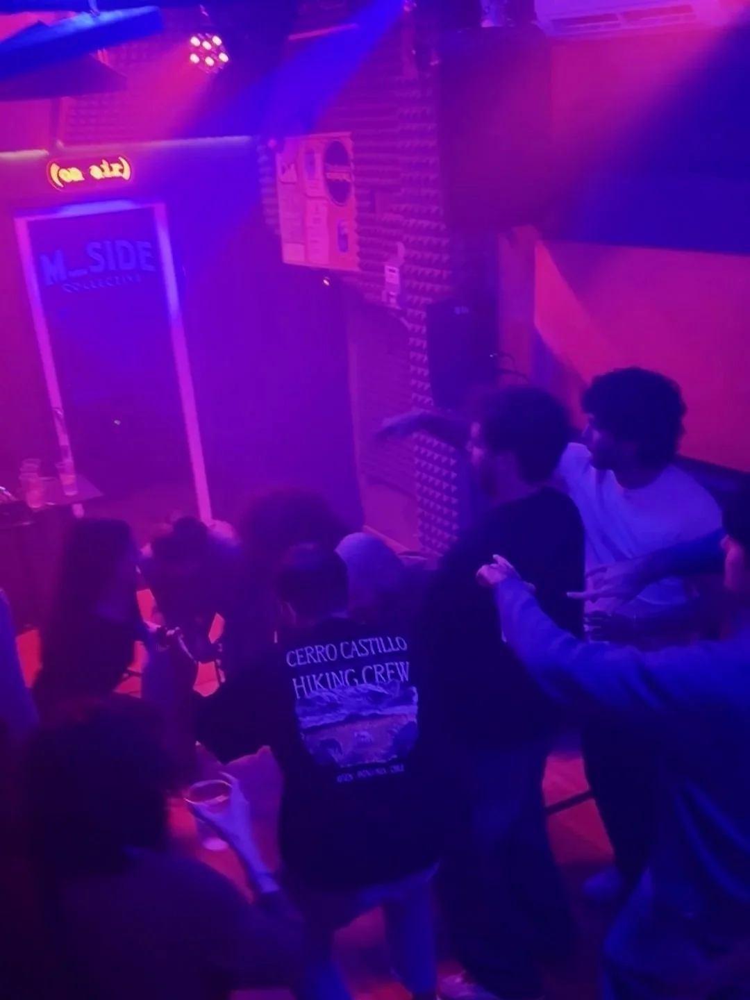 A room with purple and pink lighting, people seated and standing, some holding drinks, and a sign that reads 'M_SIDE'.
