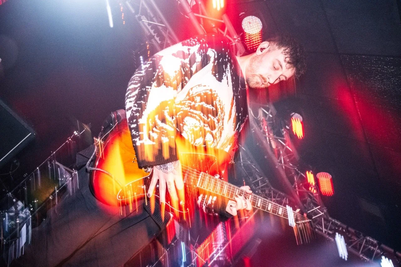 Tropic Gold. A musician playing an acoustic guitar on stage with red and orange lights and stage equipment visible in the background.