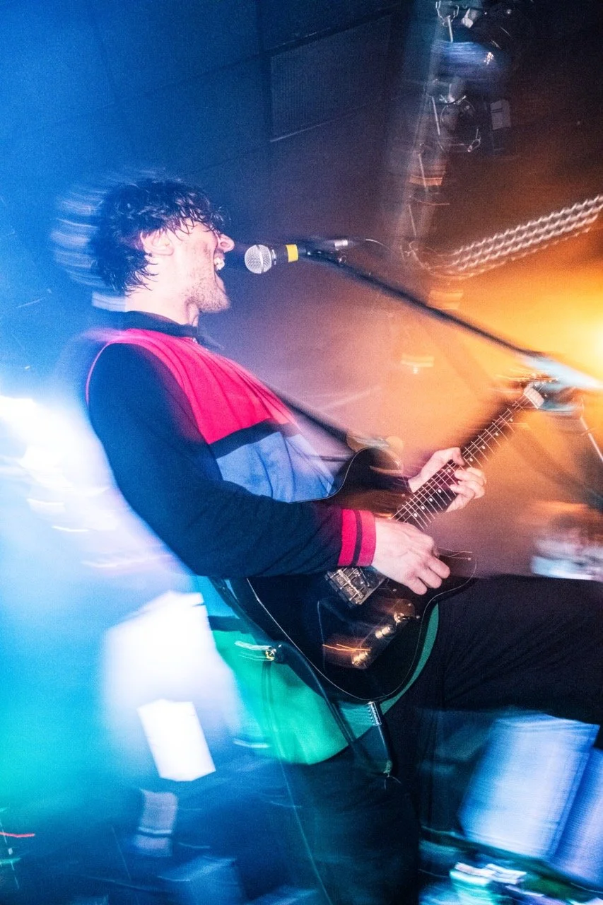 Luke Caley. unpeople. A person with wet hair singing into a microphone while playing a guitar on stage in a vibrant, colorful lighting setting.