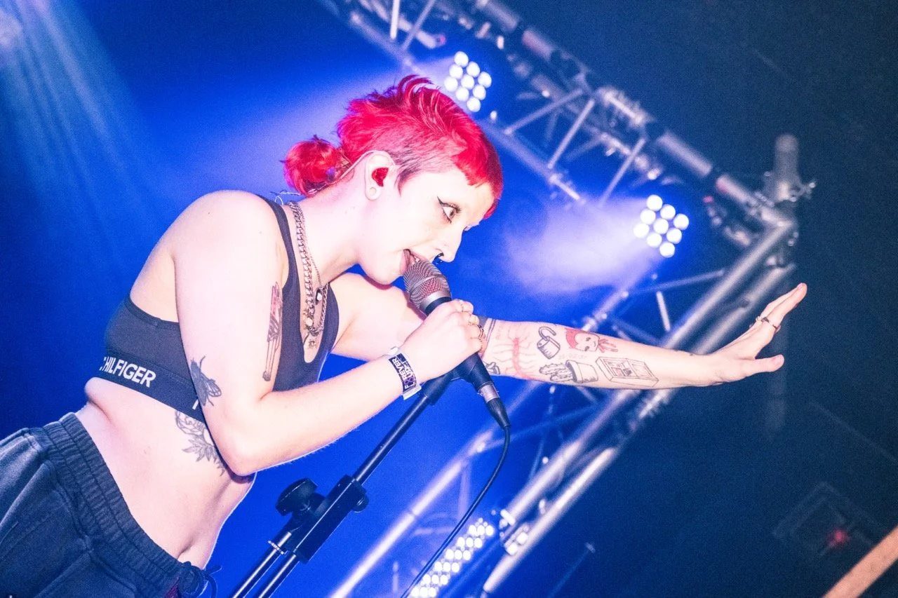 Cody Frost. A female singer with short red hair performing on stage, holding a microphone with one hand and extending her arm toward the audience. She has visible tattoos on her arm and torso, with stage lighting in the background.