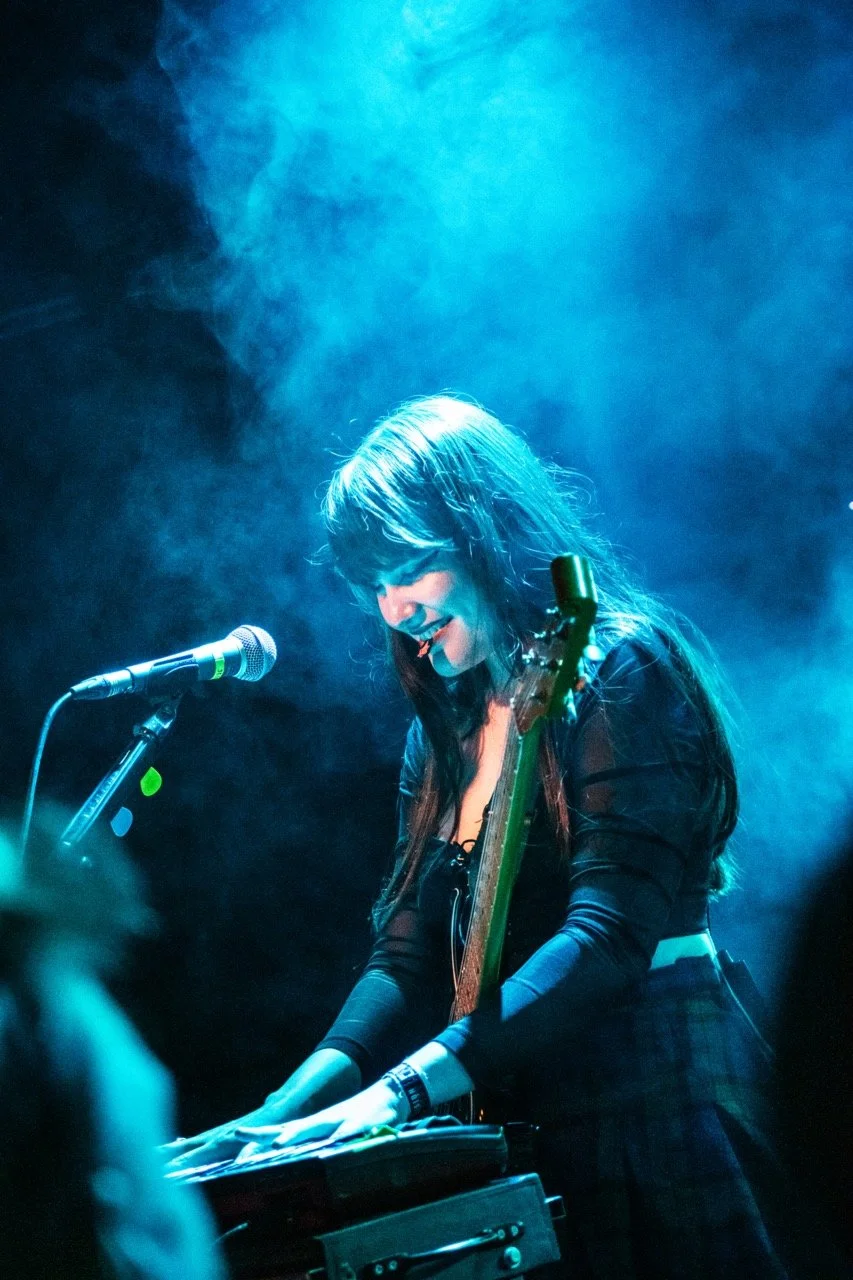 Calva Louise. Female musician playing keyboard with a guitar strap over shoulder on stage, illuminated by blue stage lights and fog.