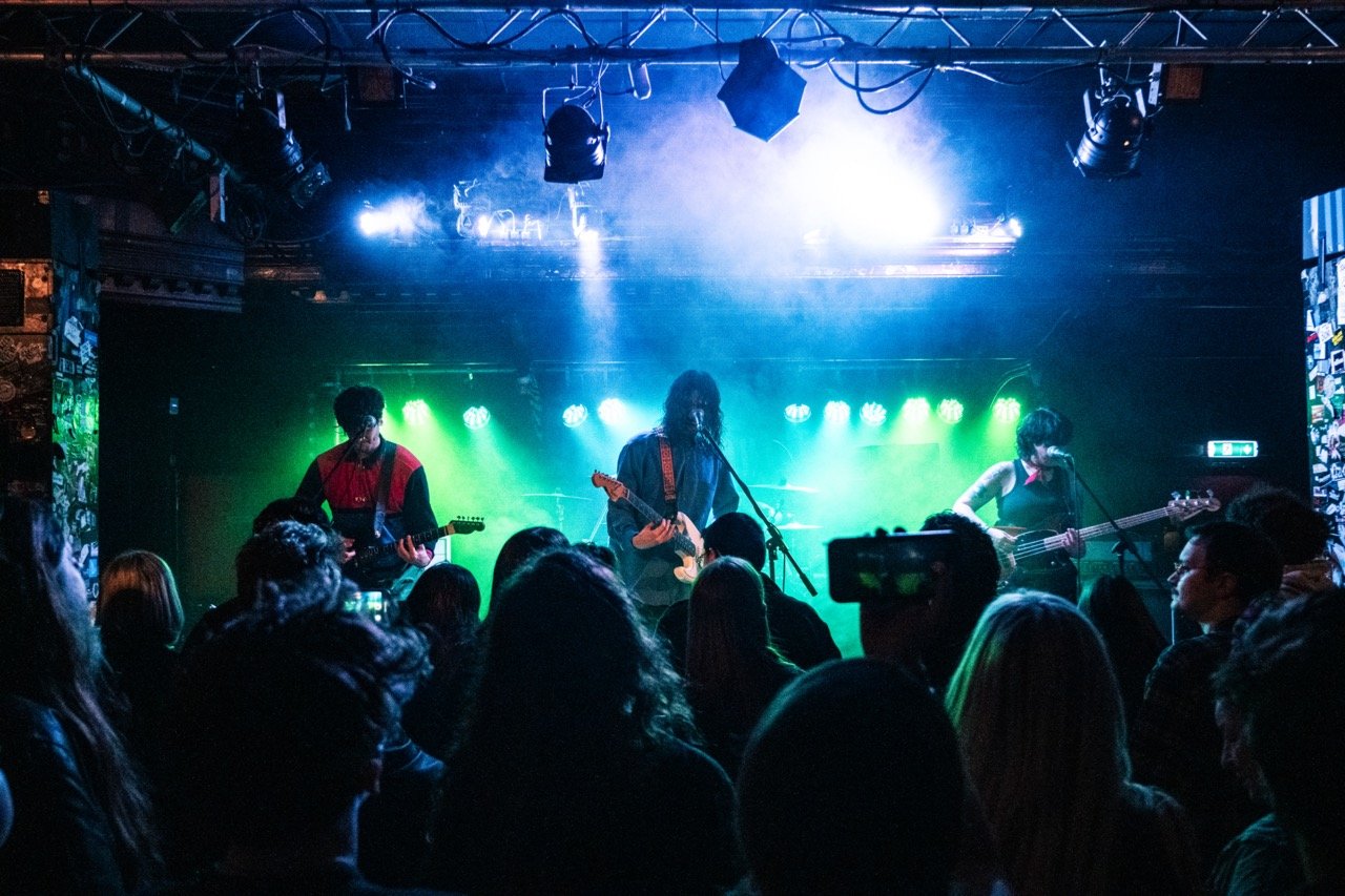 unpeople. A band performing on stage with green and blue lighting, with three members playing guitars and singing into microphones, while an audience watches.