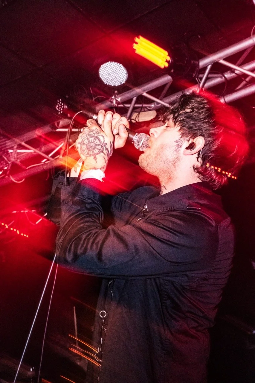Tropic Gold. A male singer with tattoos on his hand, holding a microphone with both hands, performing under red stage lights in a dark venue.