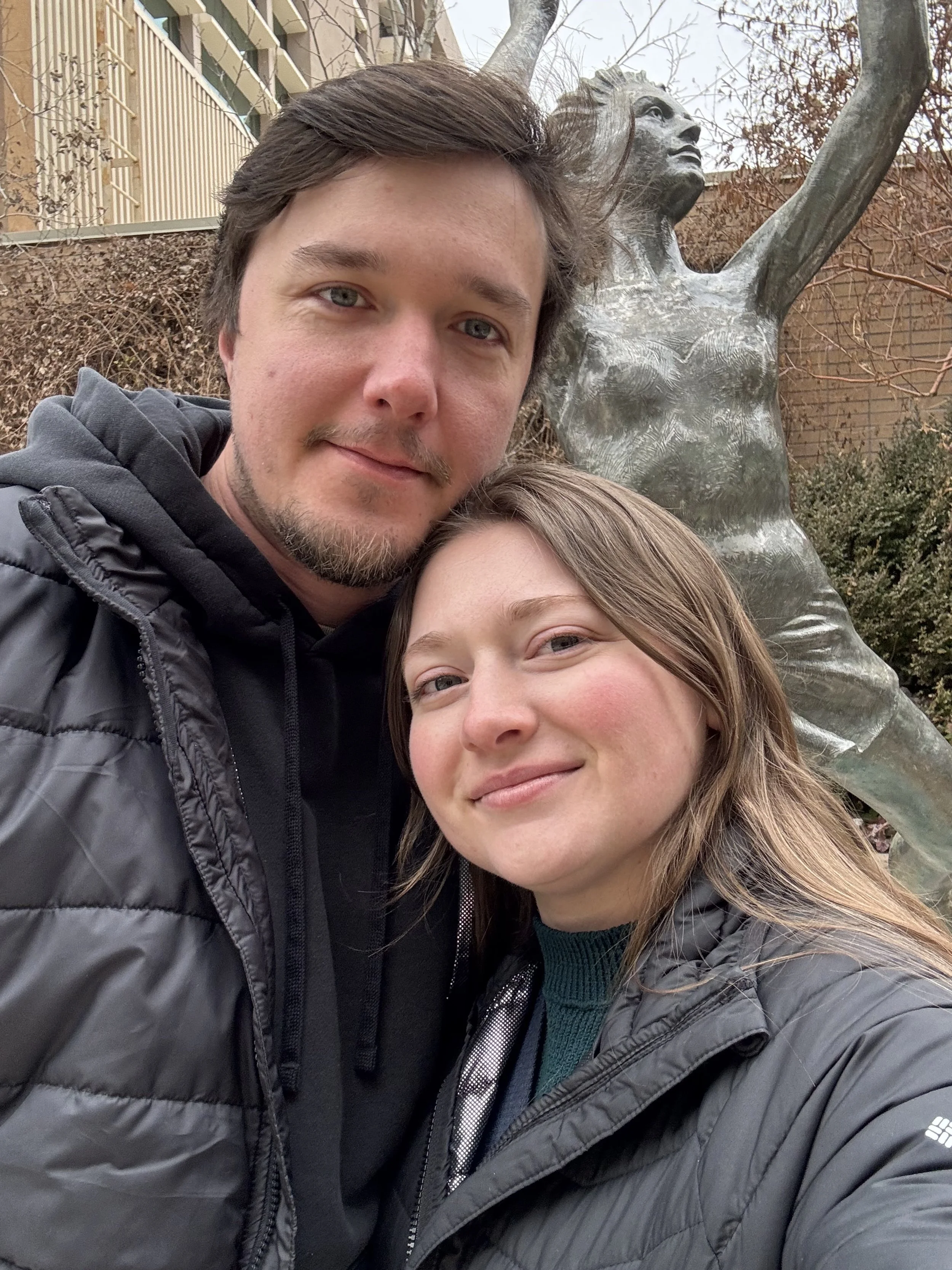 A young man and woman take a selfie outdoors in front of a bronze sculpture of a woman with her arms raised. They are both wearing jackets, and there are trees and a building in the background.