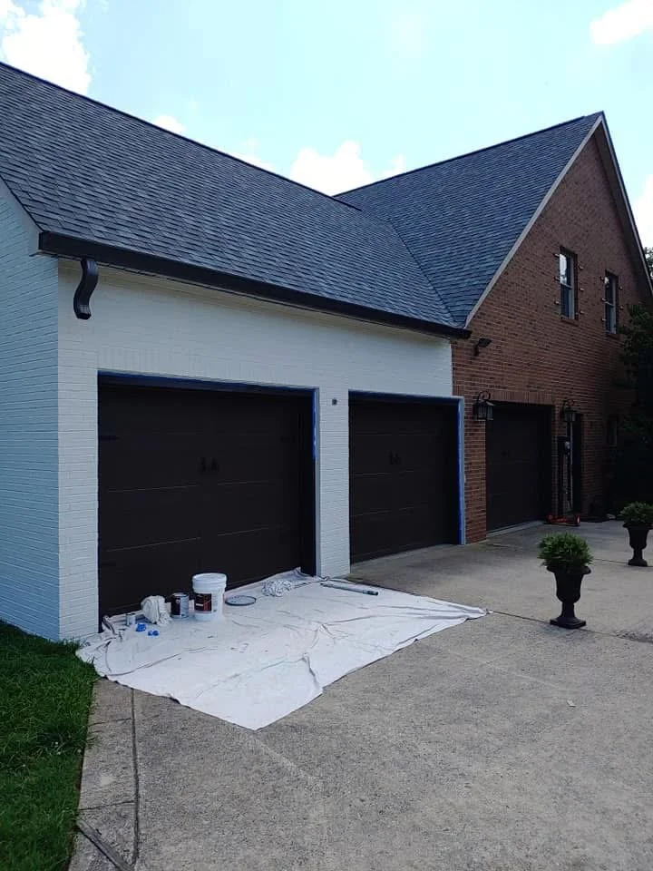 Garage doors being painted with black paint, painting supplies on a sheet on the driveway, and potted plants on either side of the driveway.