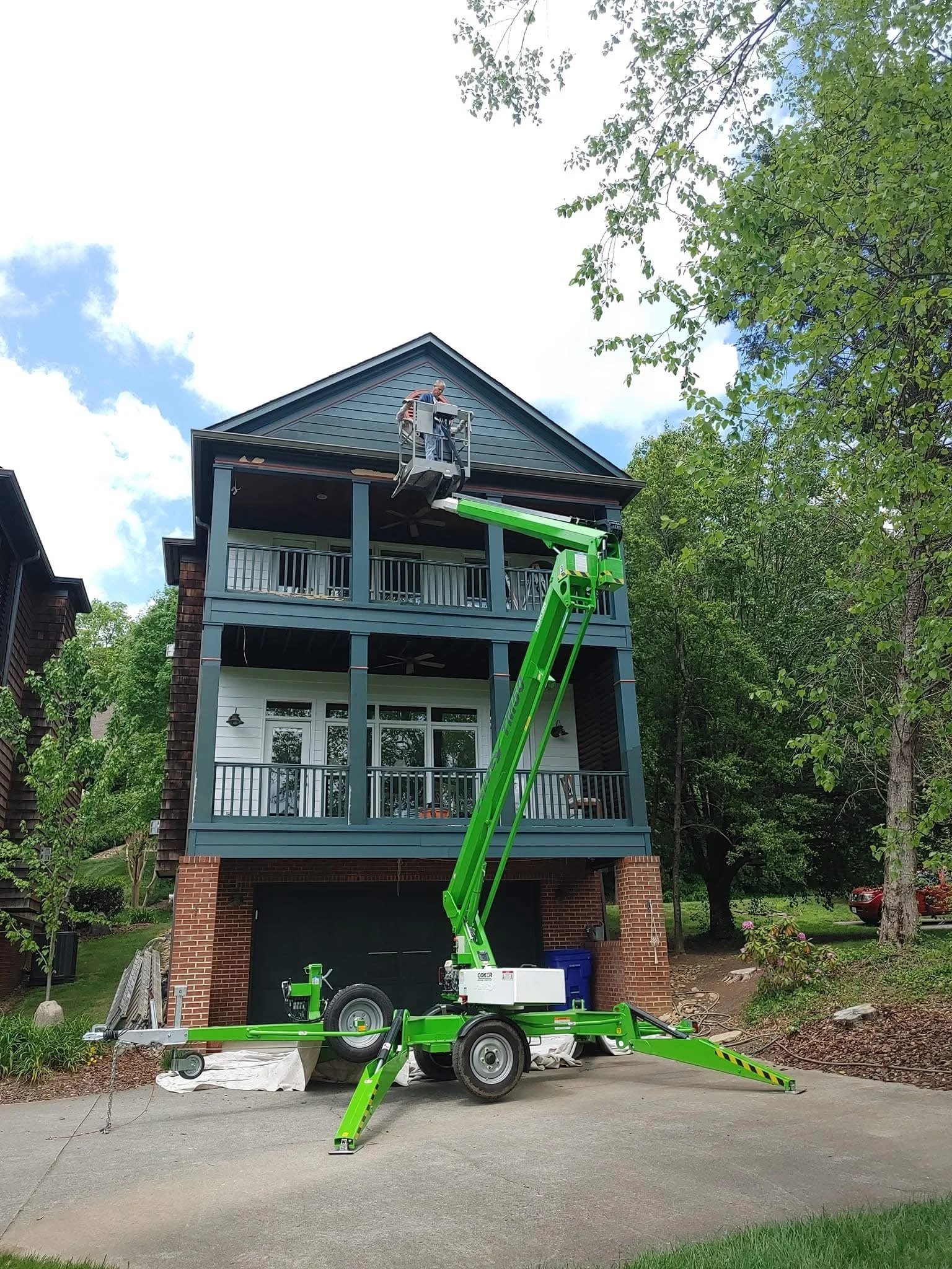 A person on a lift working on the exterior of a multi-story house with blue siding, brick foundation, and balconies. The lift is green and parked on a driveway, with trees and a cloudy sky in the background.