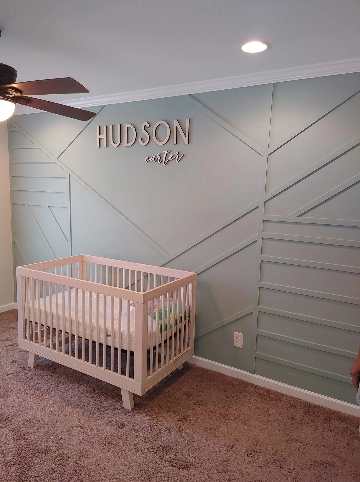 A nursery corner with a light blue wall. The wall has a geometric design with diagonal lines. The name 'HUDSON' is displayed in large letters, and the name 'carter' is below in a script font. A white baby crib with a mattress and some bedding is positioned against the wall. There is a ceiling fan with a light and wooden blades visible at the top left corner.