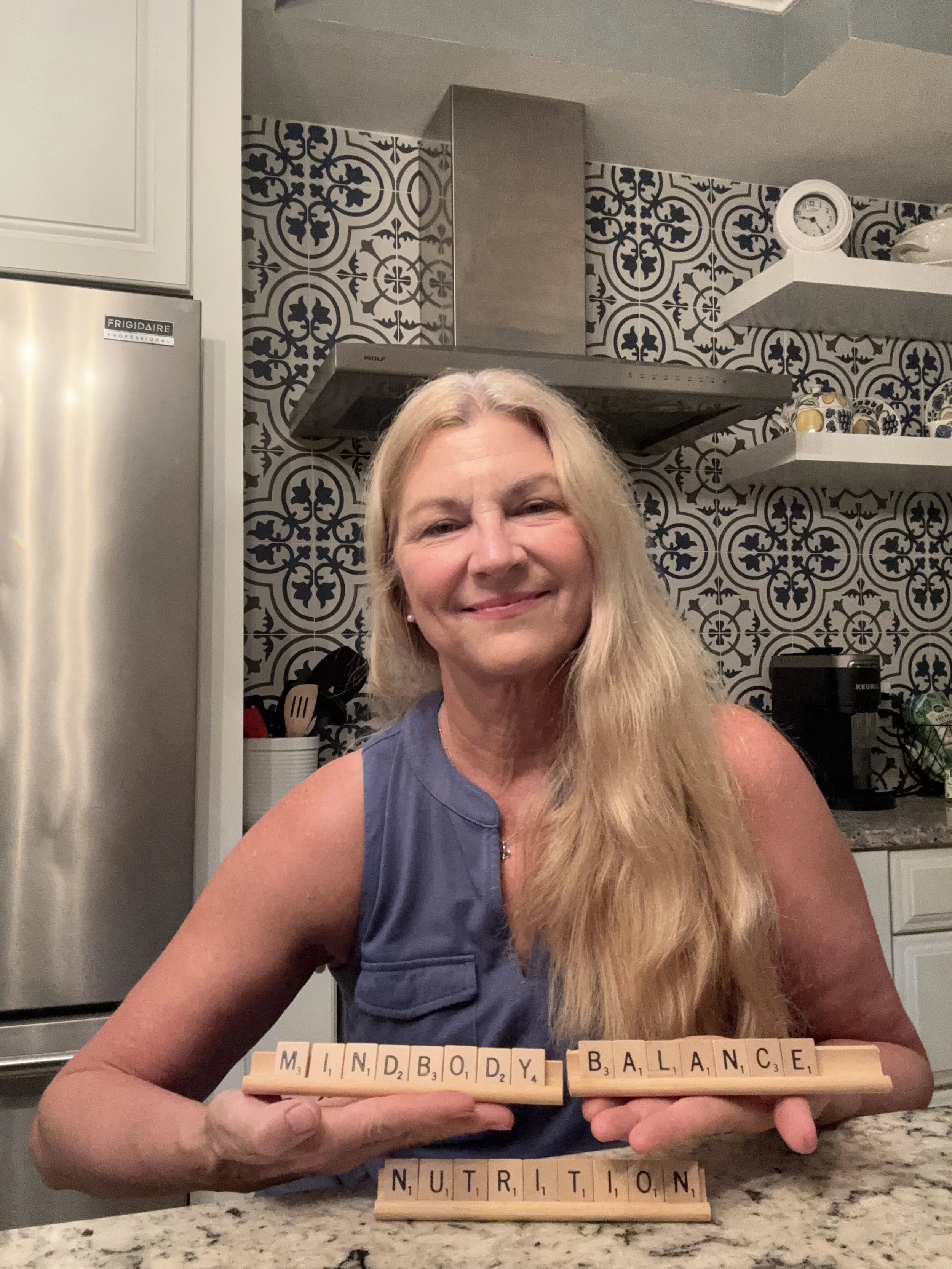 A woman with long blonde hair smiling and holding Scrabble tiles that spell out 'MIND BODY BALANCE NUTRITION' in a modern kitchen with black and white patterned tiles on the wall, open shelves with decorative items, and a refrigerator.