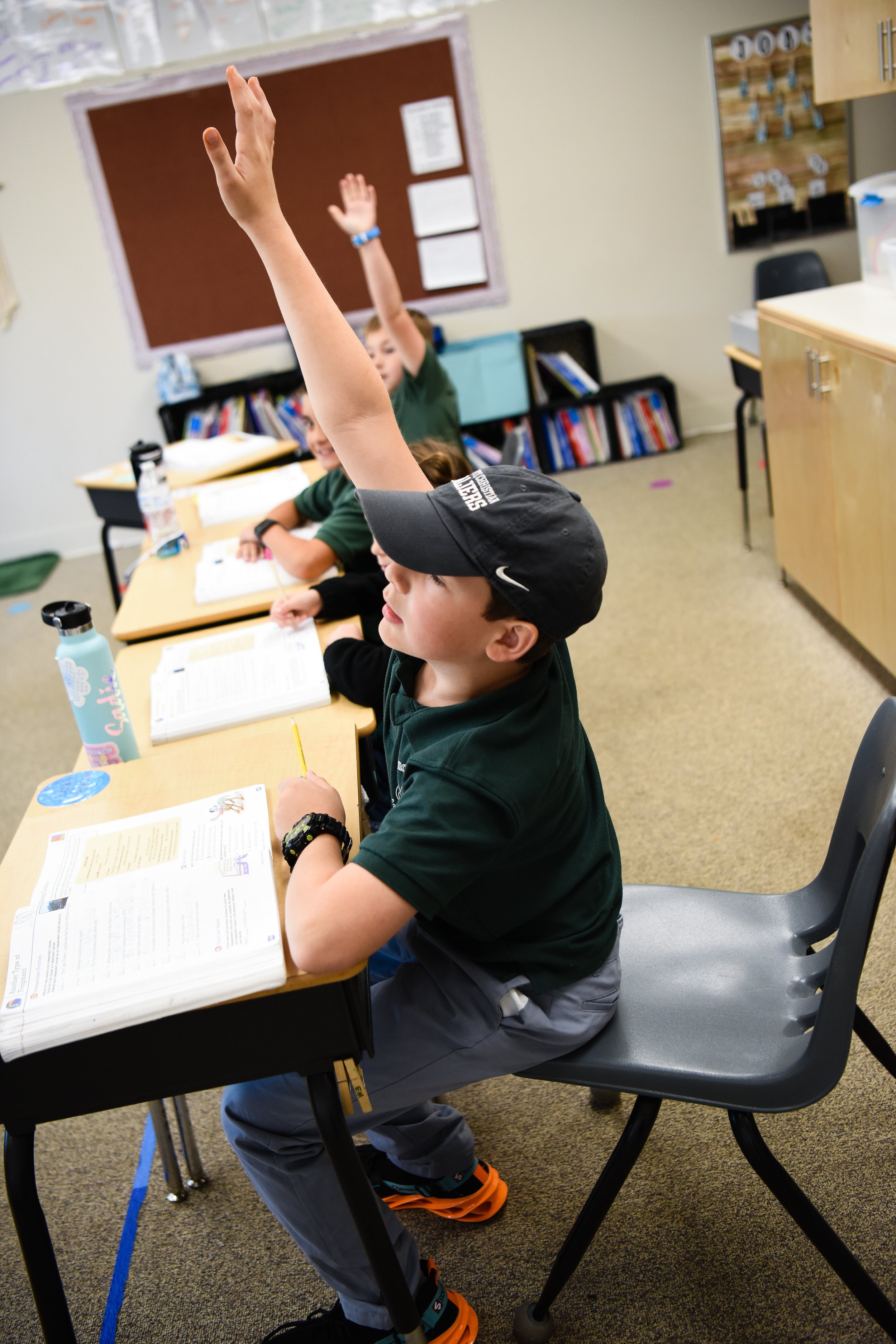 Students in a classroom raising their hands, seated at desks with books, water bottles, and stationery, with a bulletin board and classroom storage in the background.