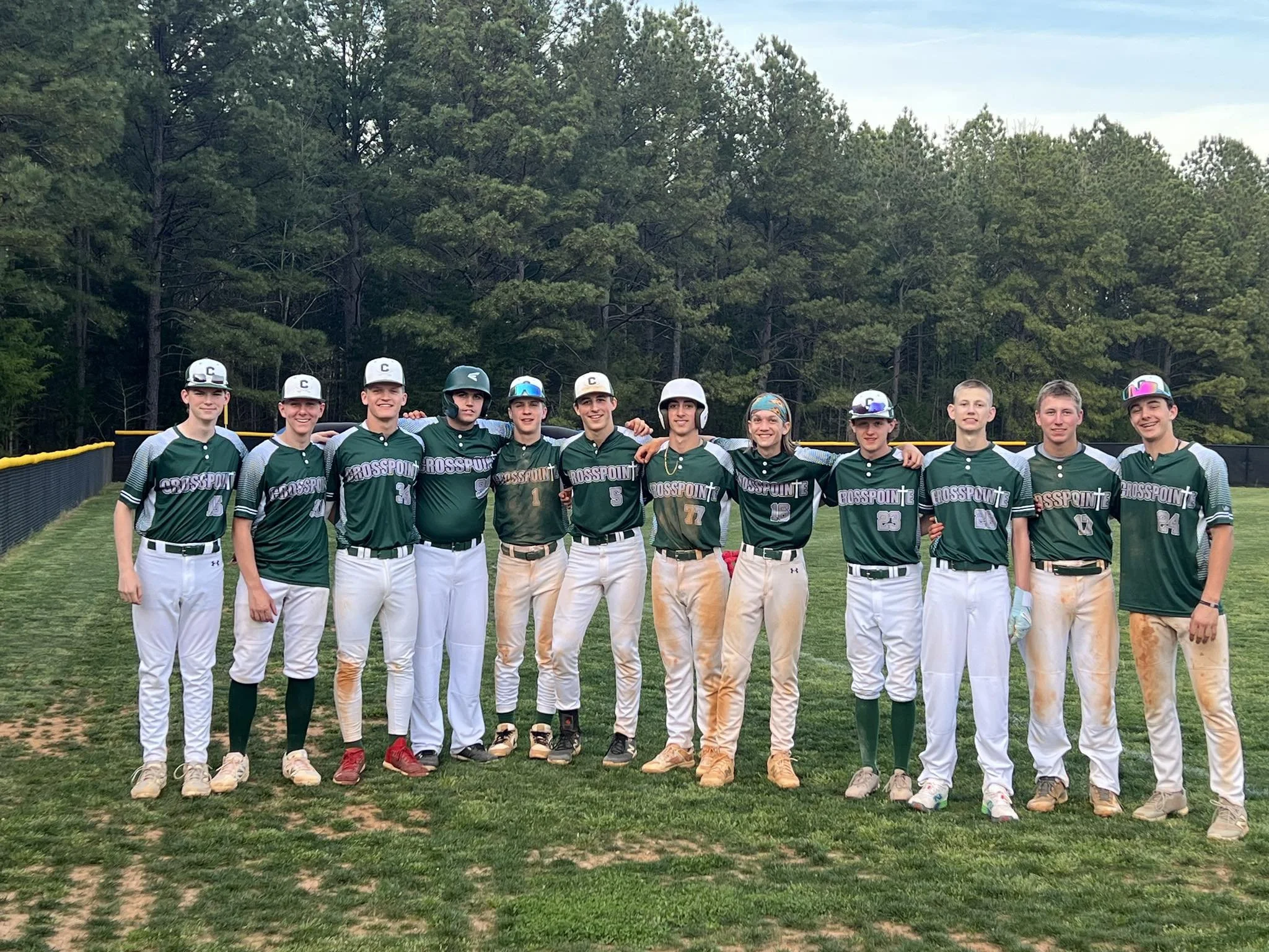 A group of eleven baseball players standing on a field with trees in the background, posing for a team photo. They are wearing green and white uniforms with 'CrossPoint' written on the front, some wearing helmets or caps.