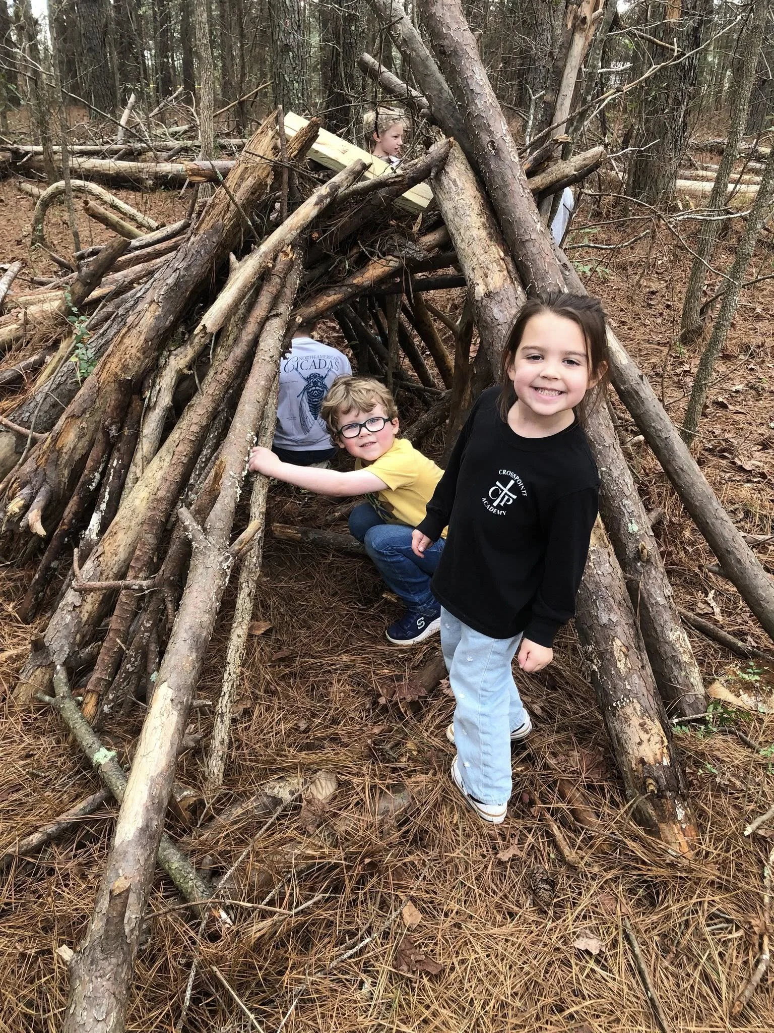 Three children playing inside a makeshift tent built from fallen branches and logs in a wooded area, with one girl smiling at the camera in the foreground.