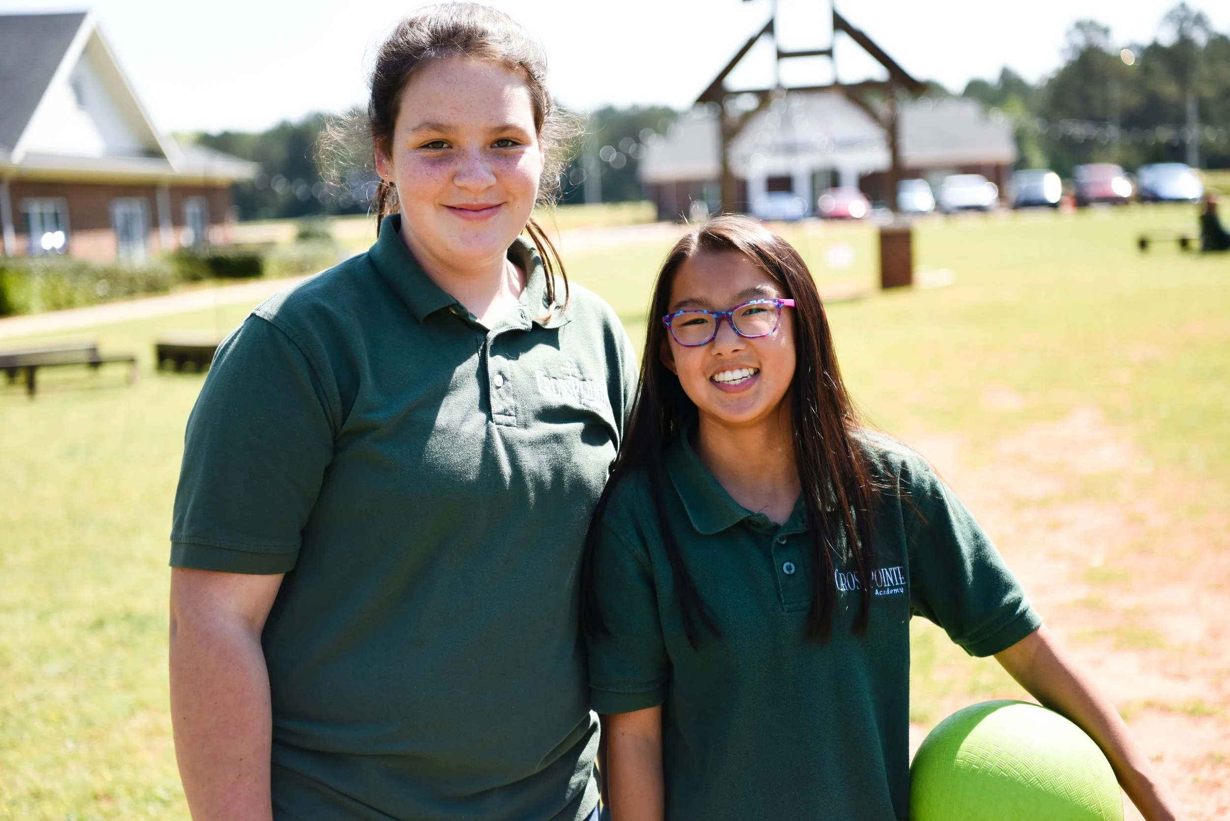 Two girls wearing green polo shirts standing outdoors in a grassy area, one holding a green ball, with buildings and parked cars in the background on a sunny day.