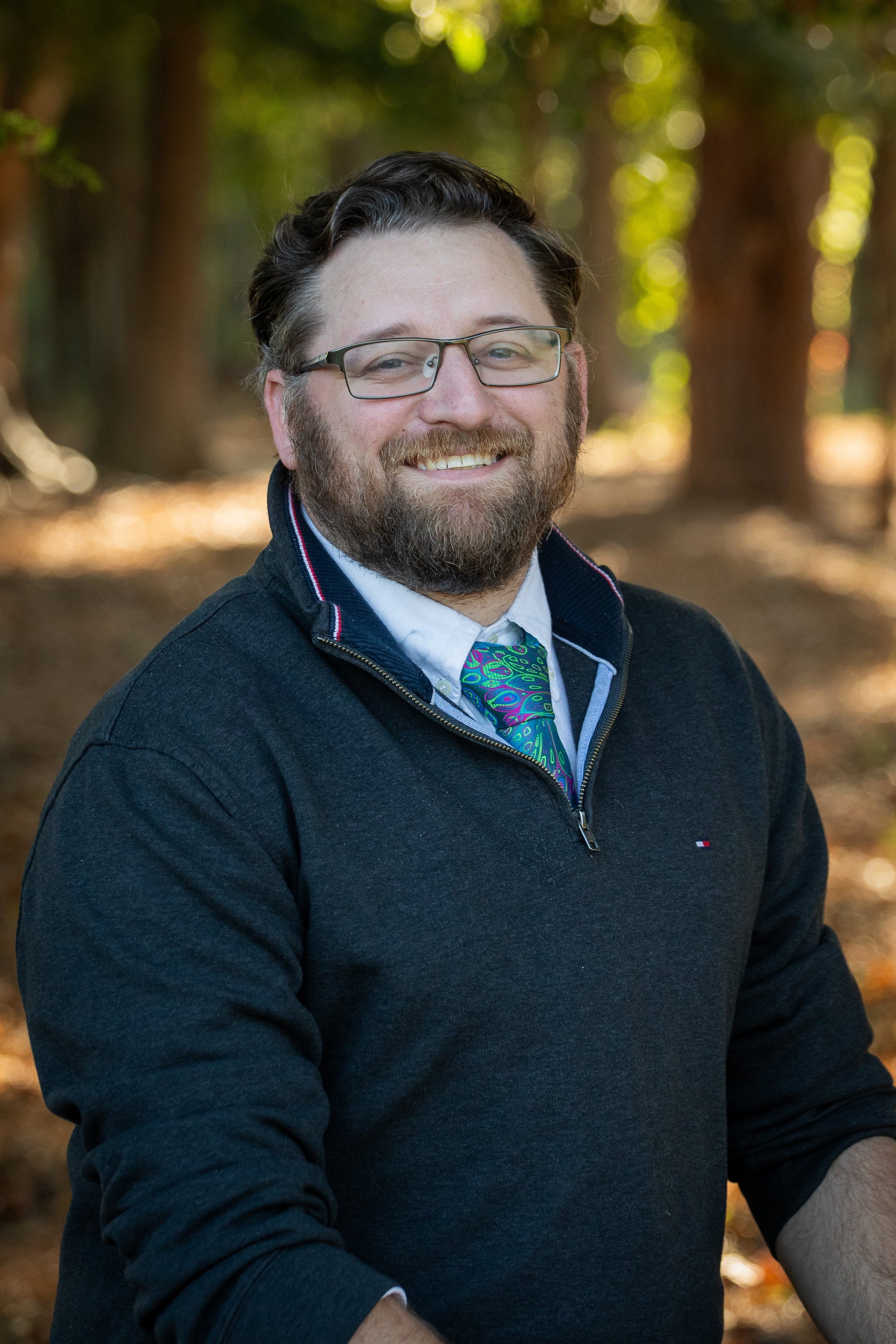 Portrait of a man with glasses and a beard, smiling, outdoors with trees and sunlight in the background.