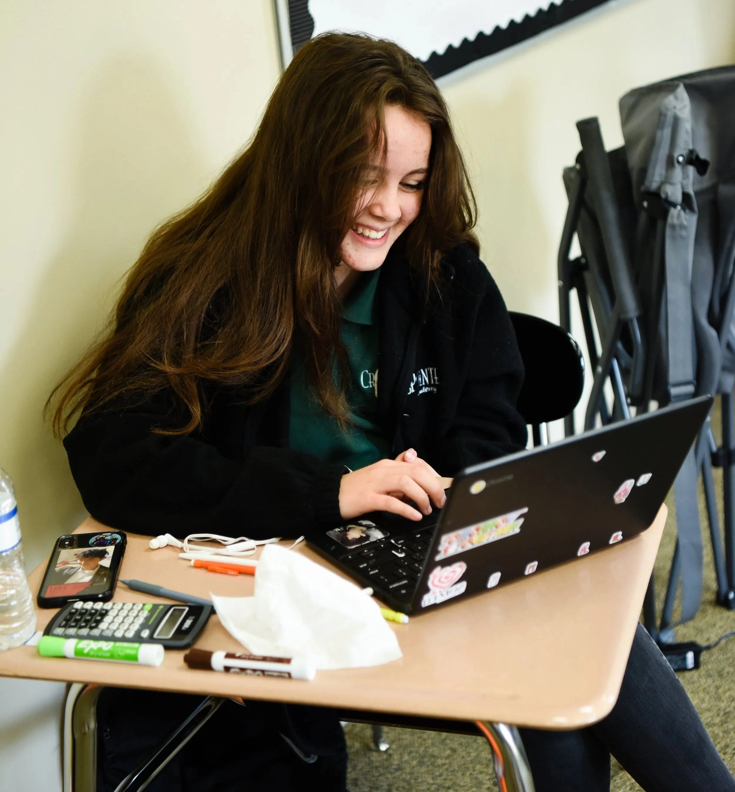 A young woman with long brown hair smiling while using a laptop on a classroom desk, surrounded by school supplies and a water bottle, with a stroller in the background.