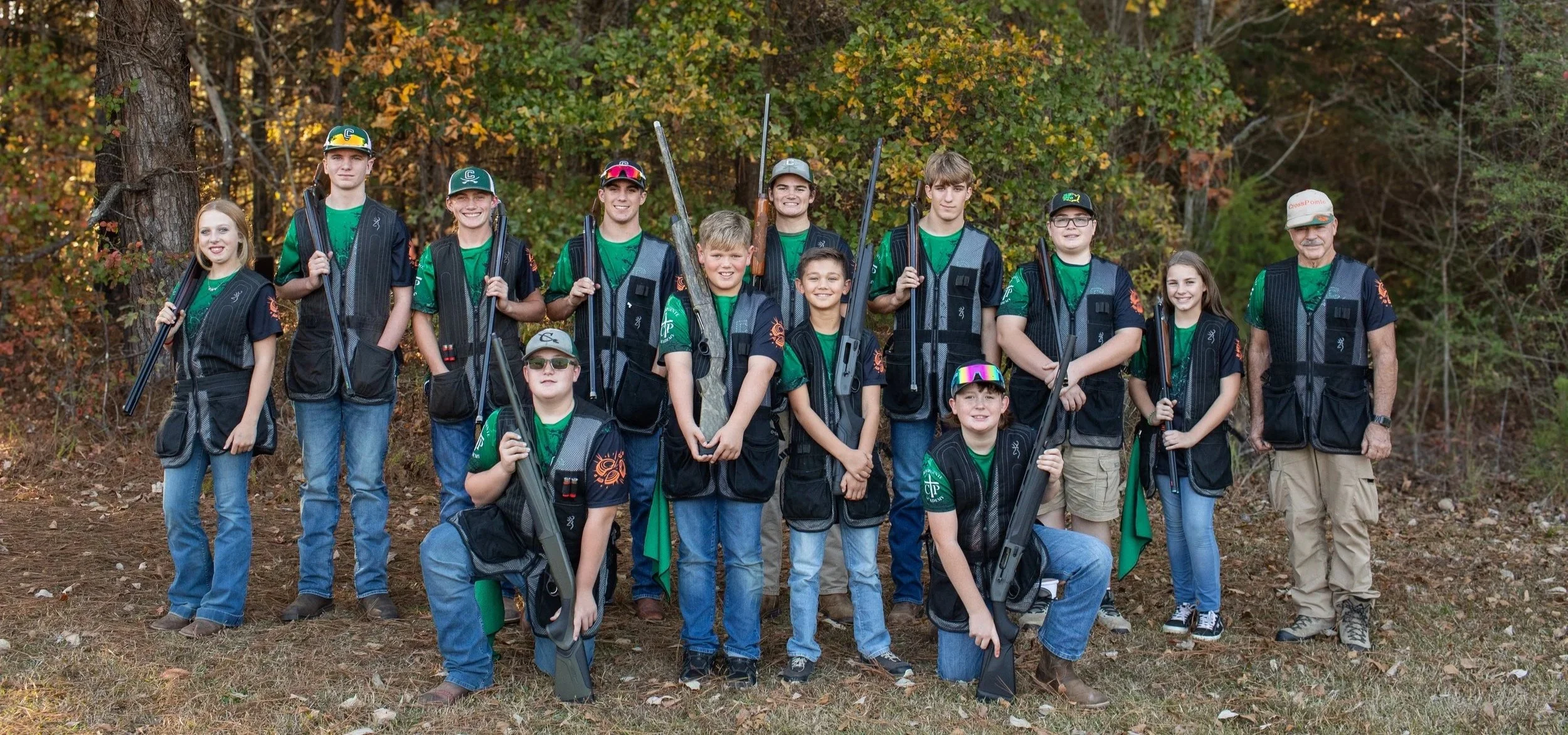 Group of young Boy Scouts and their adult leader in an outdoor wooded area, dressed in green uniforms and black vests, holding shotguns for a hunting activity.
