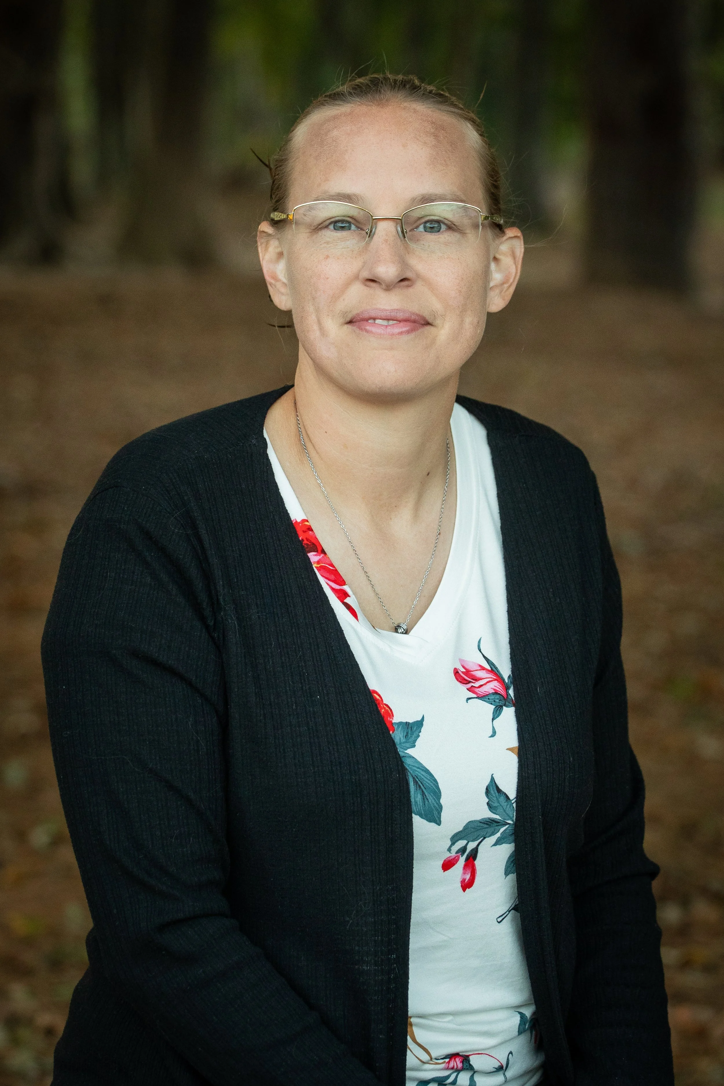 A woman with glasses and light reddish hair in a bun, wearing a black cardigan over a white shirt with a floral pattern, standing outdoors in a wooded area.
