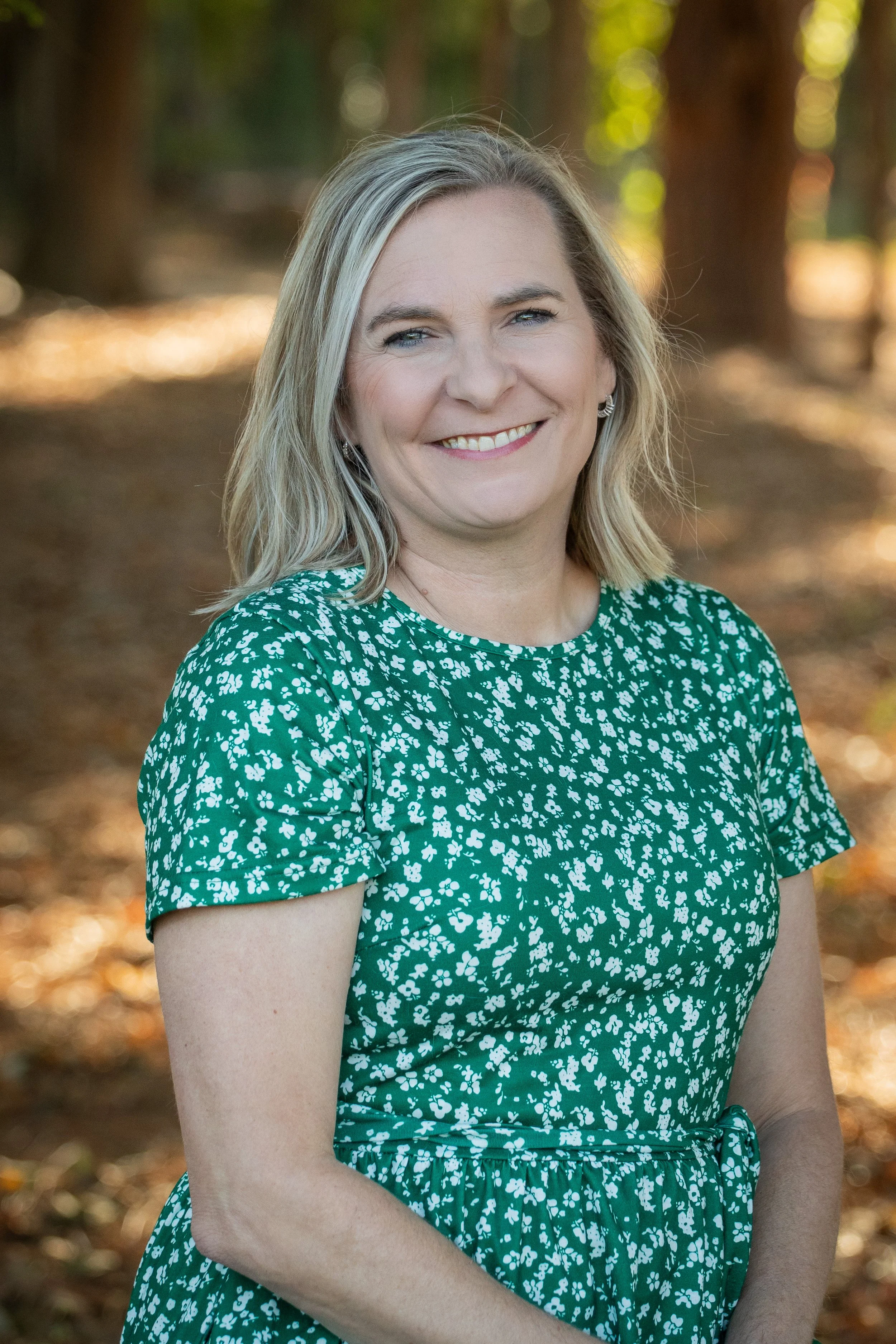 A woman with shoulder-length blonde hair, smiling, outdoors in a wooded area, wearing a green and white floral dress.