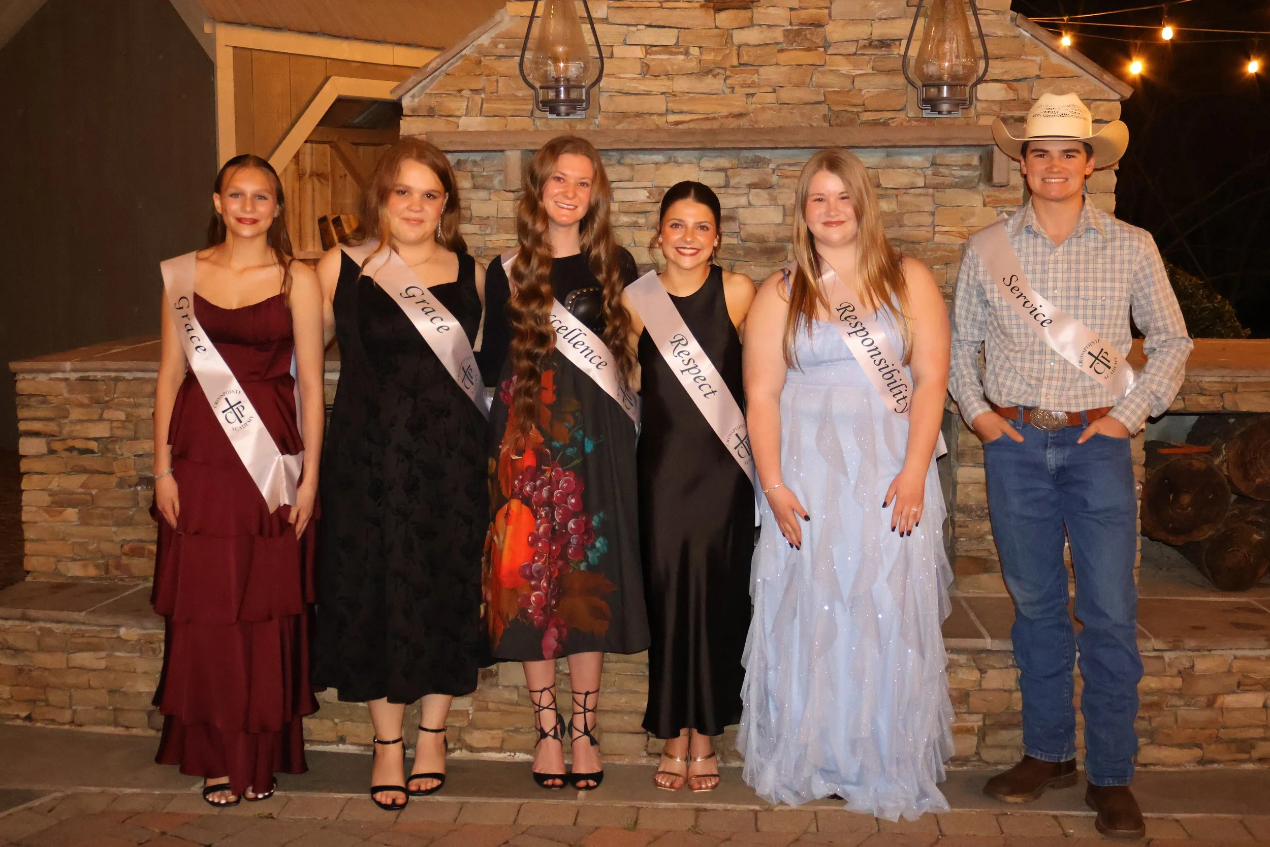 Group of six young people, five women and one man, posing in front of a brick fireplace with hanging lanterns. They are dressed in formal attire, and each wears a sash with words like 'Grace,' 'Faith,' 'Excellence,' 'Respect,' 'Responsibility,' and 'Service.' The man is wearing a cowboy hat and a plaid shirt.