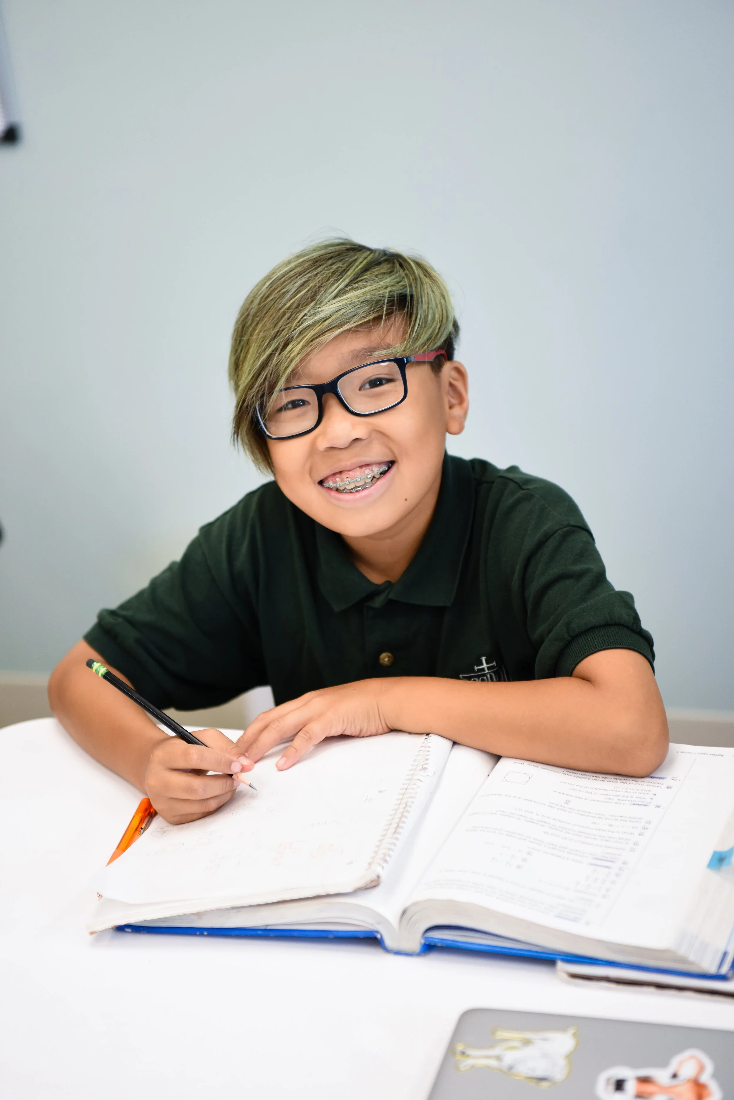 A smiling boy with glasses and braces, sitting at a table with an open book and writing materials, in a classroom setting.