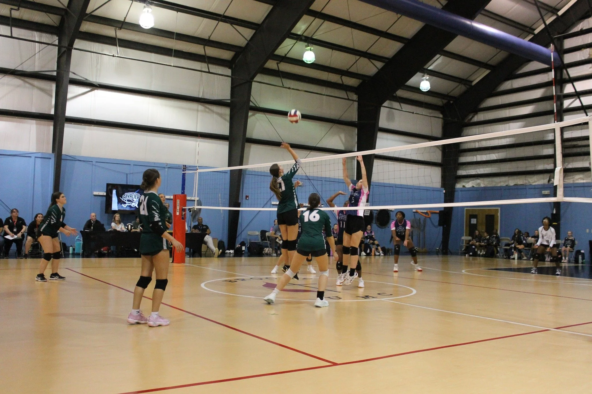 A girls' volleyball game taking place indoors, with players jumping to hit the ball over the net, while others watch and prepare to react, with spectators seated in the background.