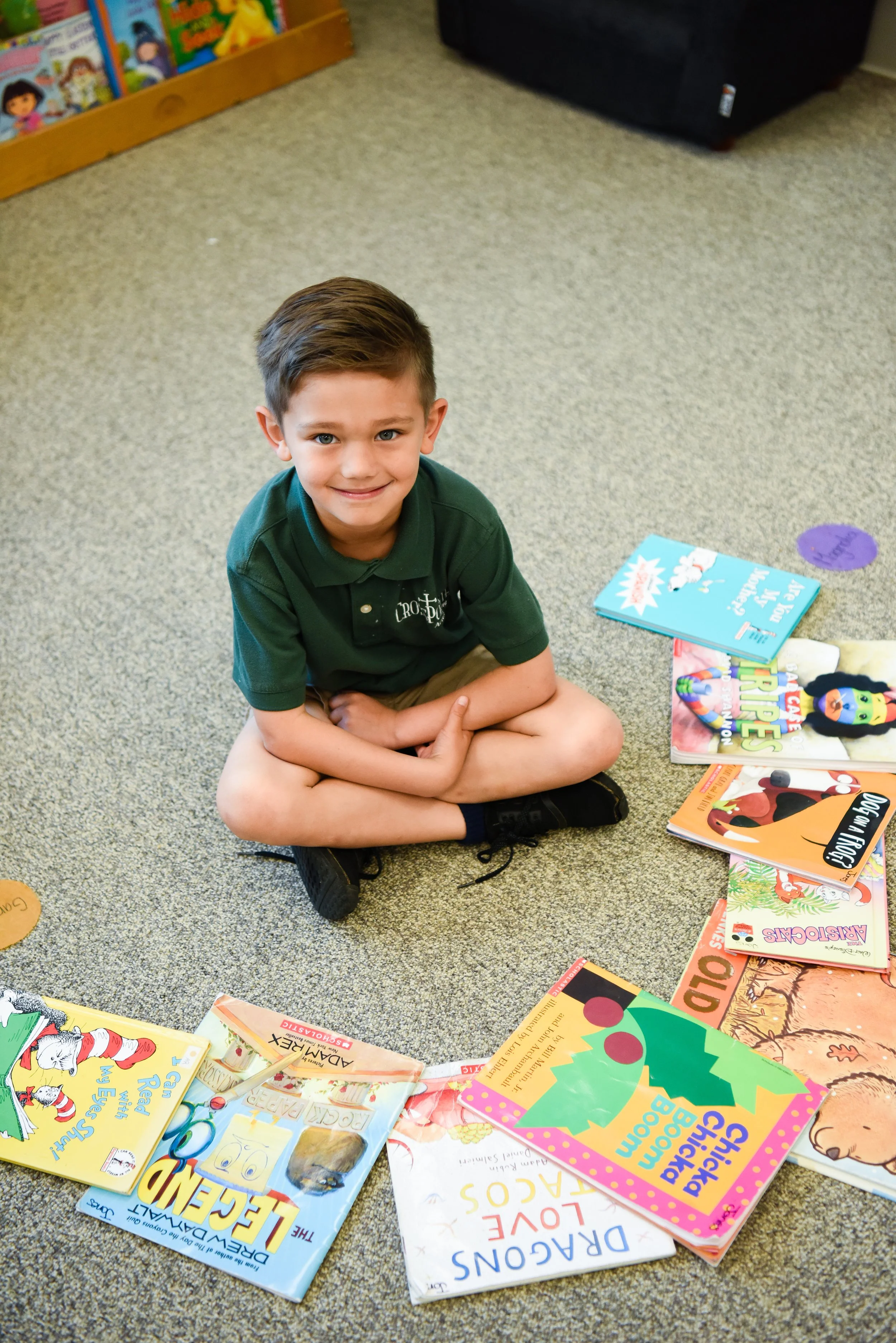 Smiling young boy with brown hair sitting cross-legged on a carpeted floor, surrounded by colorful children's books, at a library or classroom.