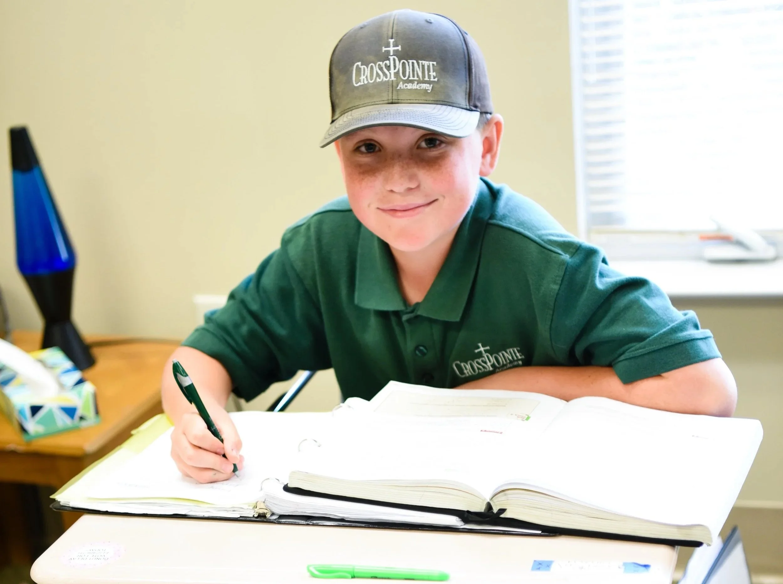 A young boy with freckles, wearing a green polo shirt and a gray cap with 'CrossPointe Academy' written on it, sitting at a desk with an open notebook and a green pen in hand, smiling at the camera.