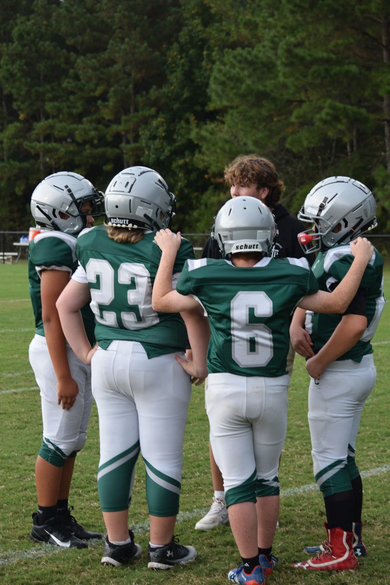 Youth football players in green jerseys and white pants huddle with a coach on a football field.