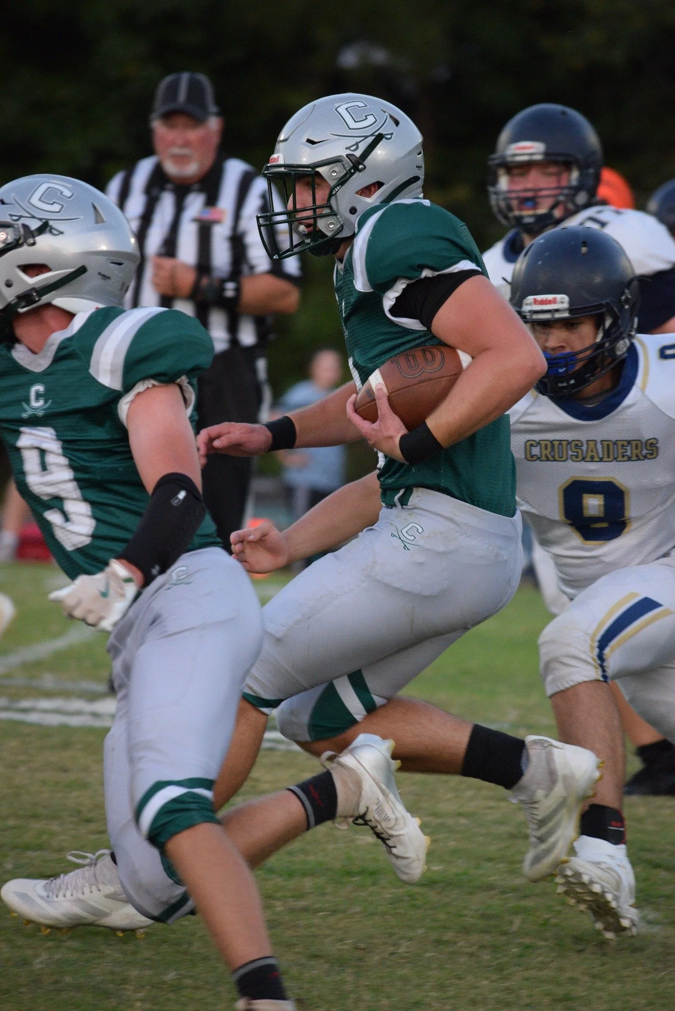 A football game showing a player in a green jersey running with the ball, tackled by a player in a white jersey. Other players and a referee are visible in the background.