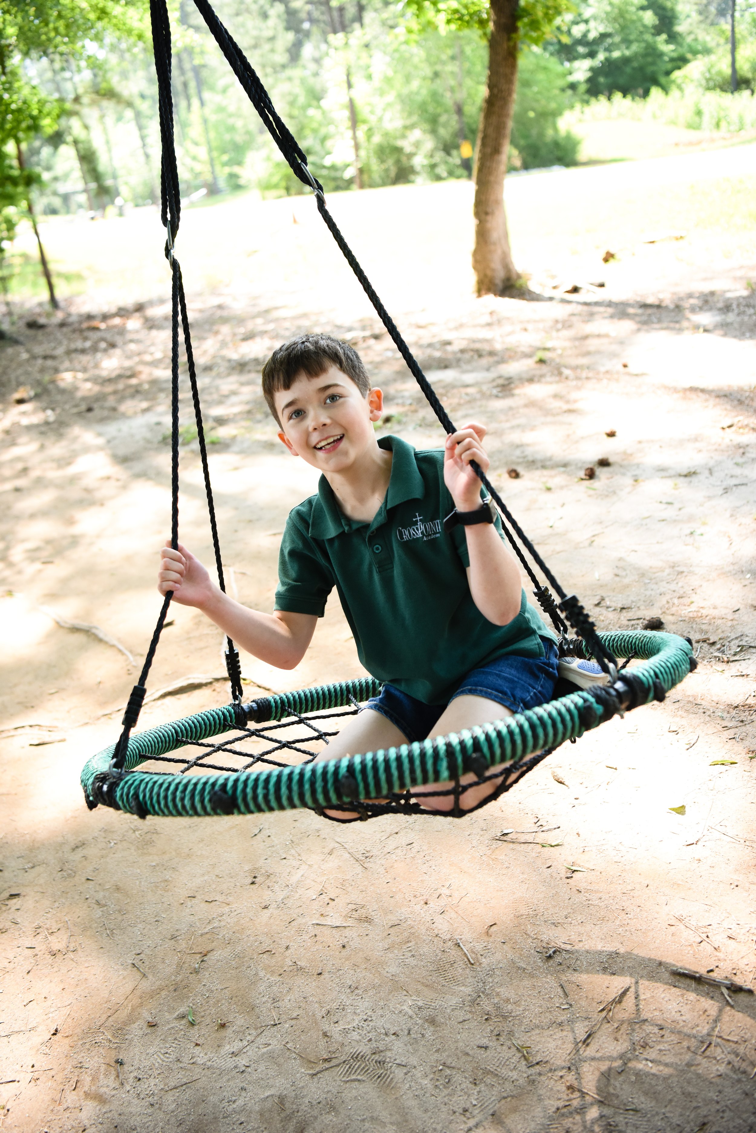 A young boy with dark hair, wearing a green polo shirt and shorts, smiling while sitting on a swing in a park with trees and greenery in the background.