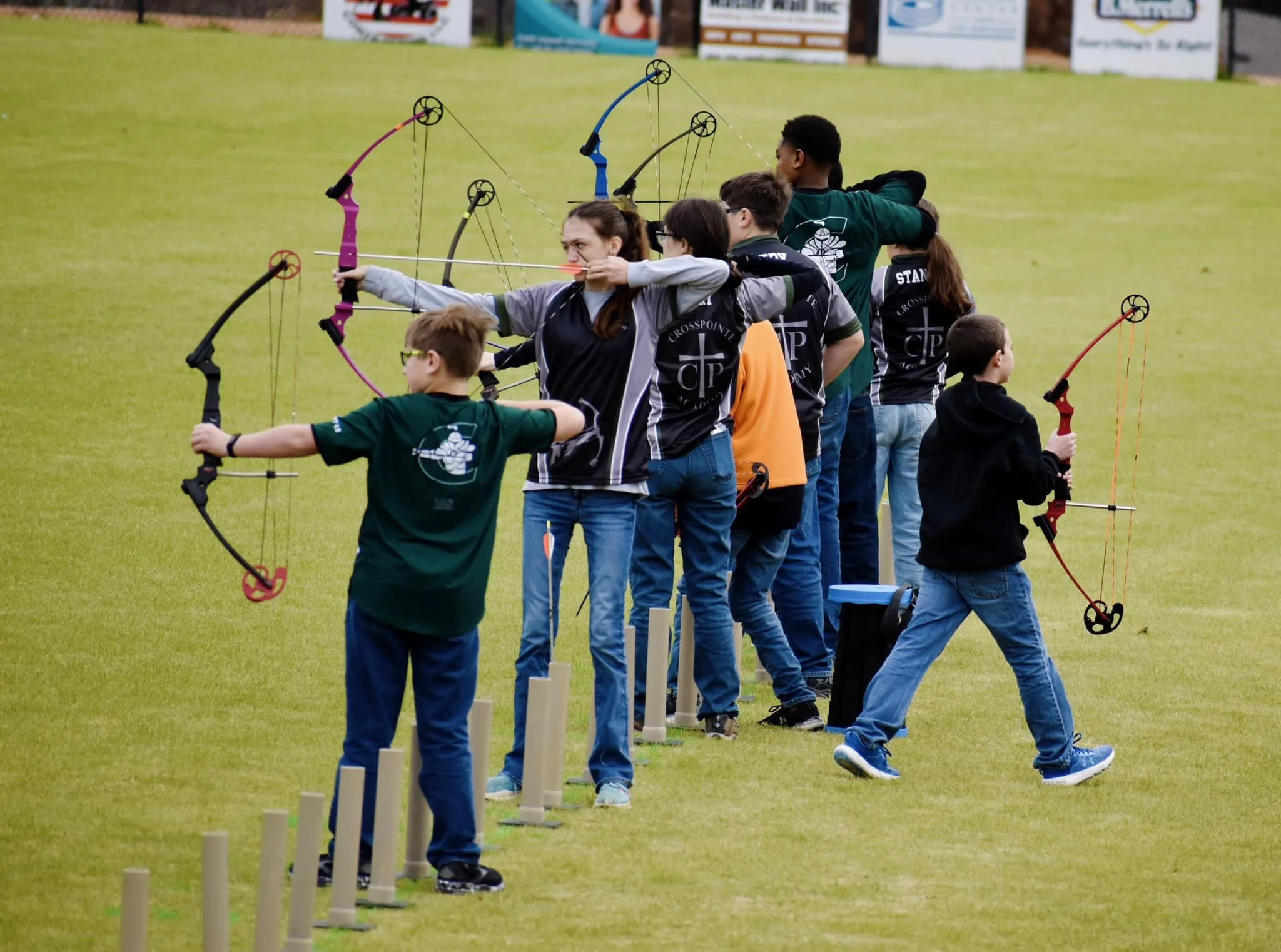 A group of children and a woman participating in an archery activity on a grassy field, aiming with bows and arrows.