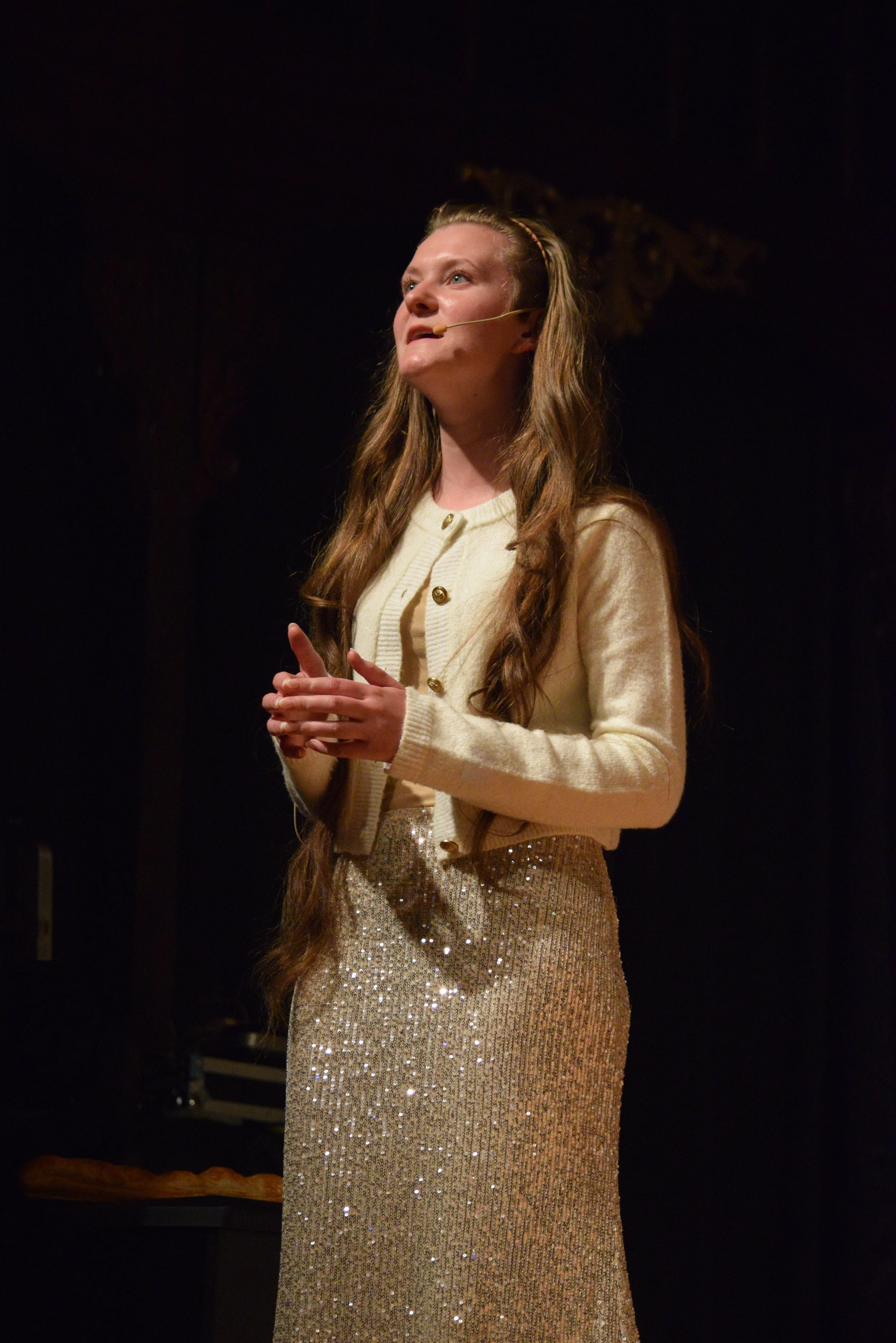 A woman with long, wavy hair standing on a stage, wearing a cream-colored cardigan and a sparkling, gold, sequin skirt, speaking into a headset microphone.