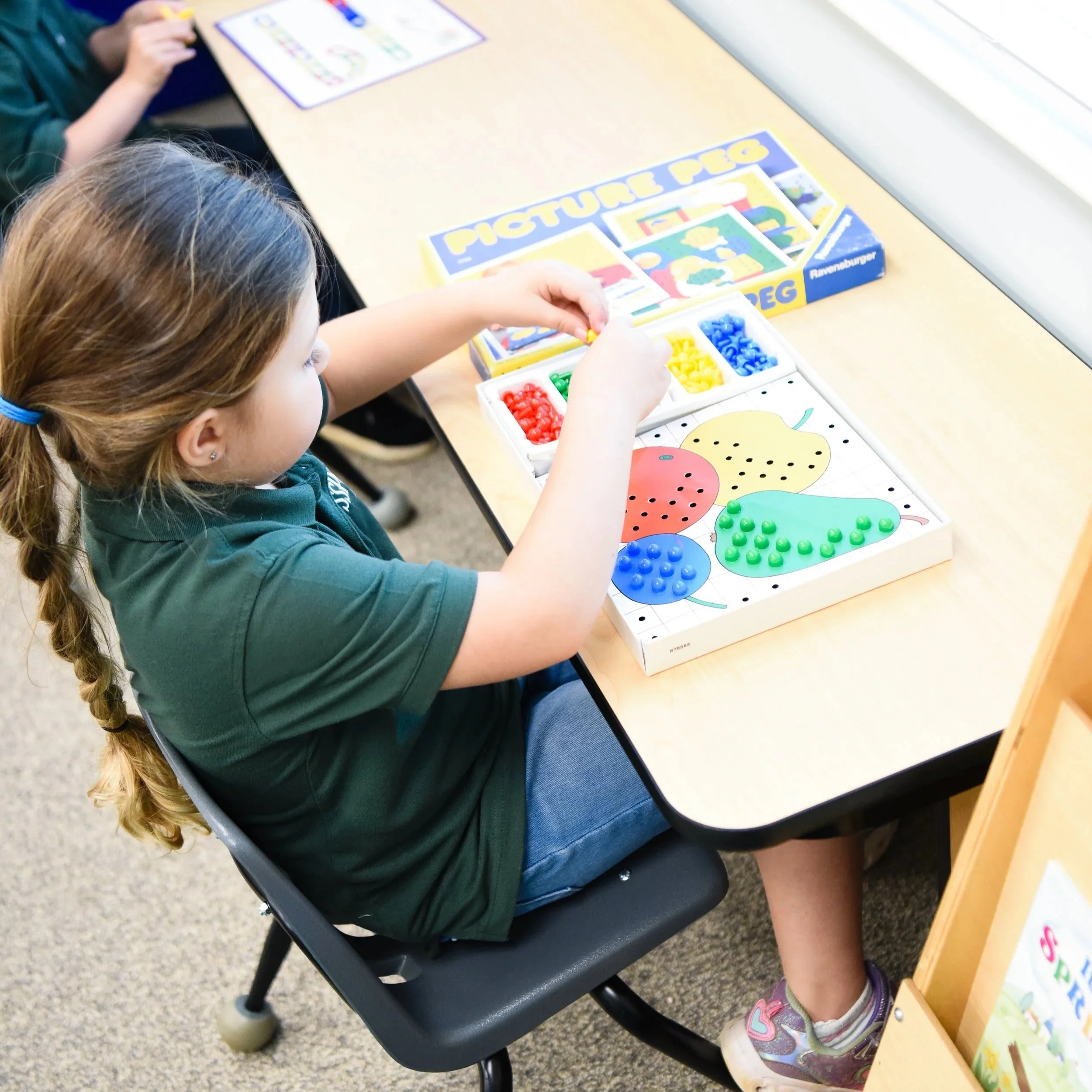 A young girl with a braid and wearing a green polo shirt and blue jeans sitting at a school desk playing a color and shape matching game with a tray of colored pegs and a board featuring different fruit shapes.