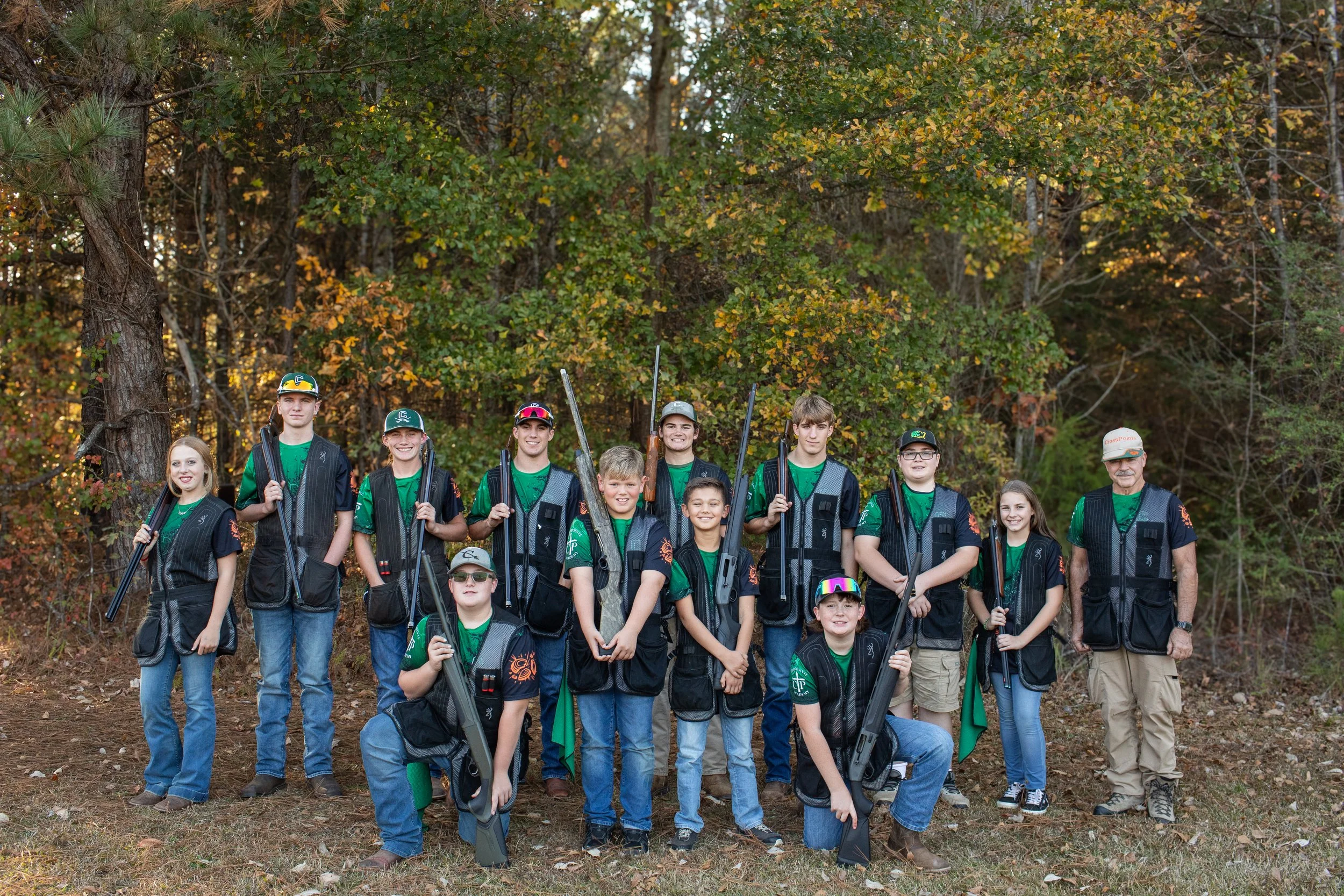 Group of young archers and their coach standing outdoors in front of trees with fall foliage, holding bows.