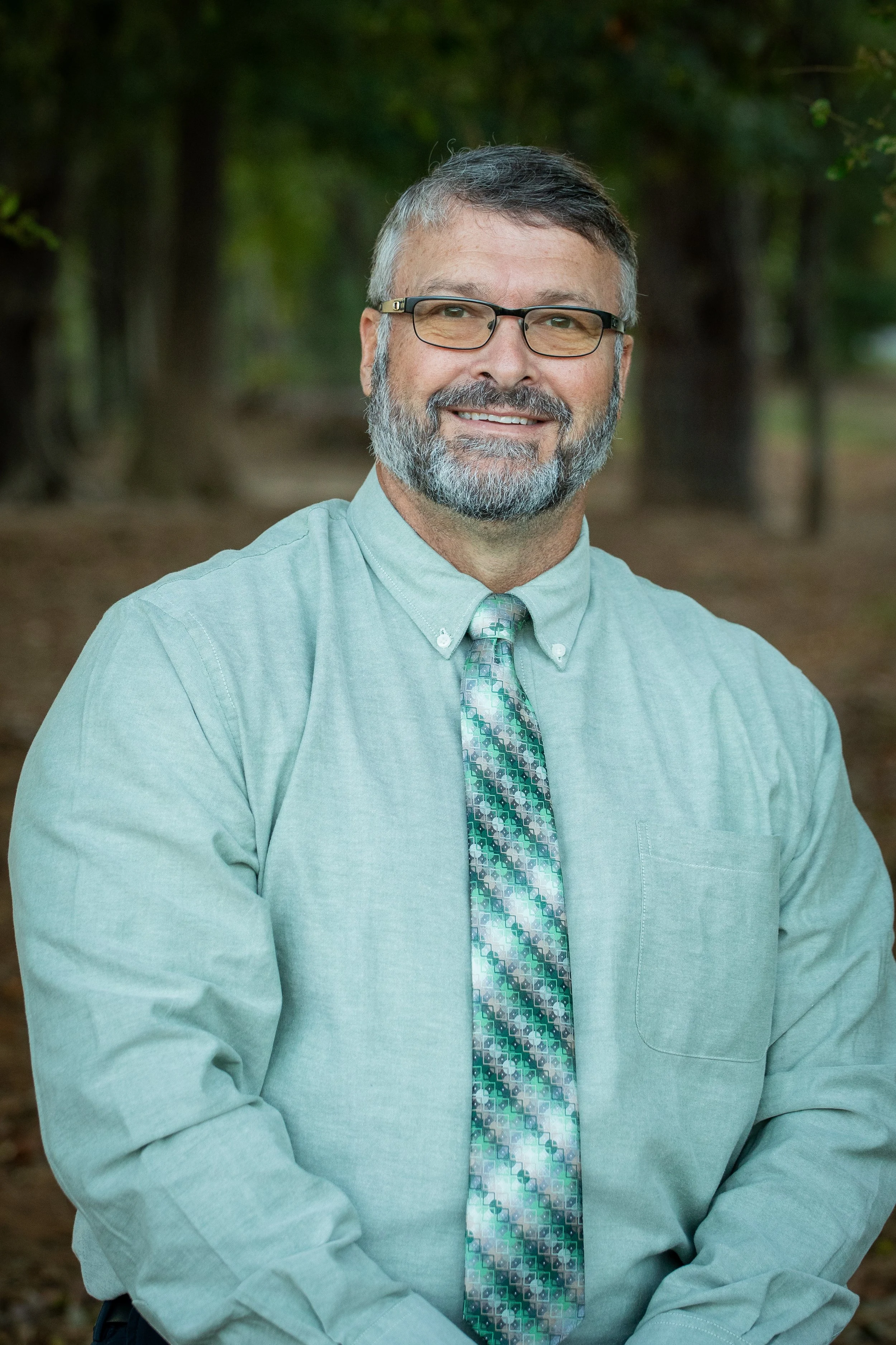 A middle-aged man with gray hair, beard, and glasses, smiling while standing outdoors in a wooded area, wearing a light green button-up shirt and a patterned tie.