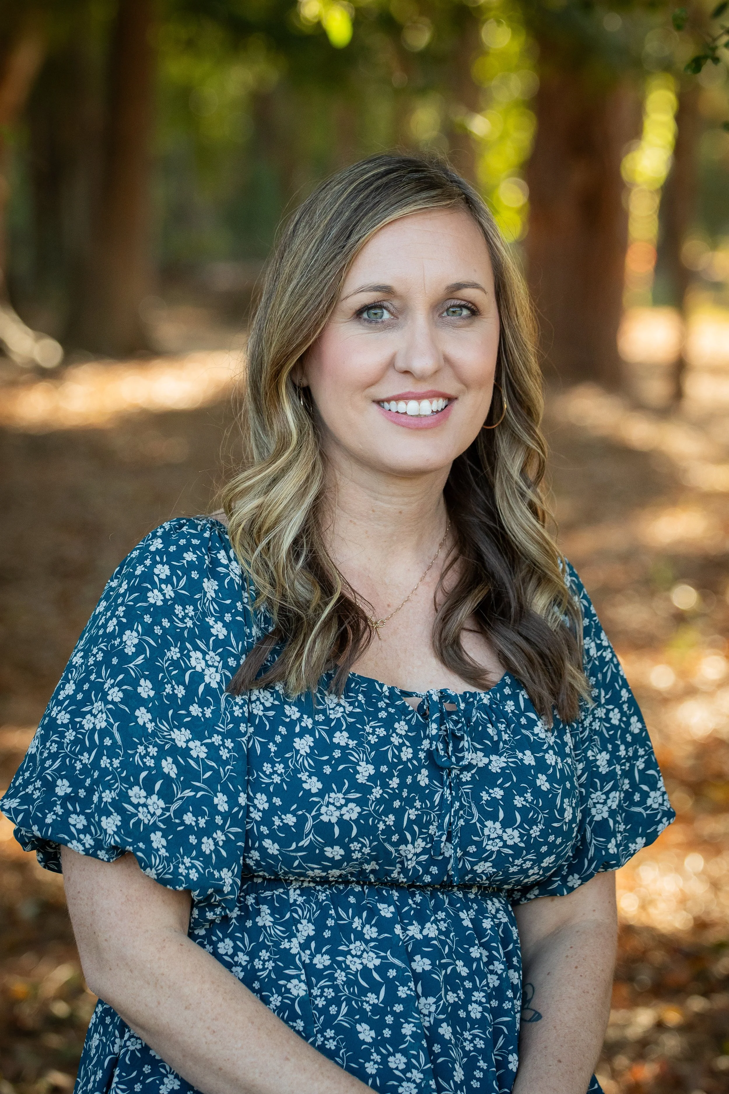A woman with long wavy hair, wearing a blue floral dress, standing outdoors in a forested area with sunlight filtering through the trees.