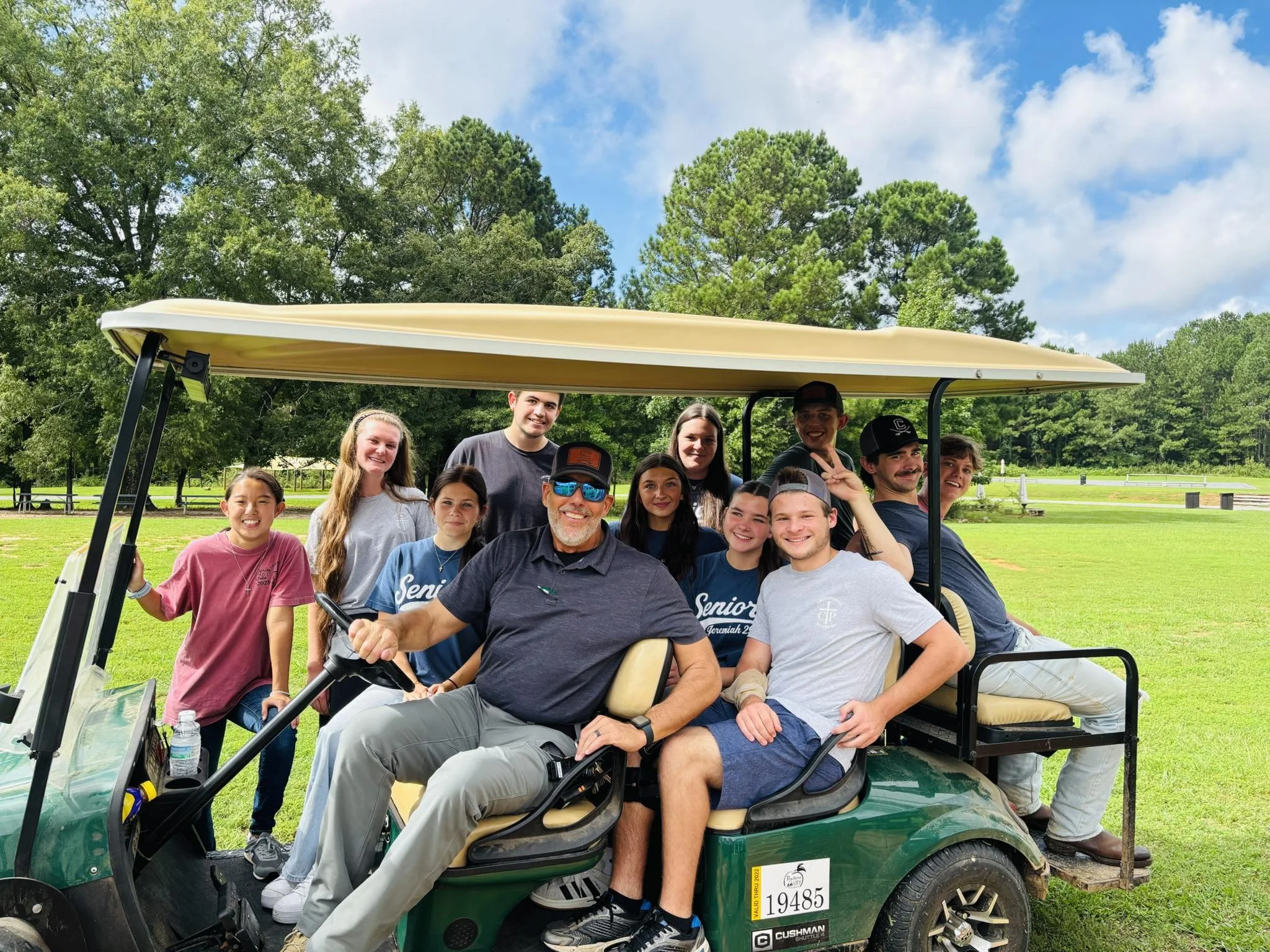 A group of young adults and one older man posing on a green golf cart in a grassy park with trees in the background. Some are smiling and making playful gestures.