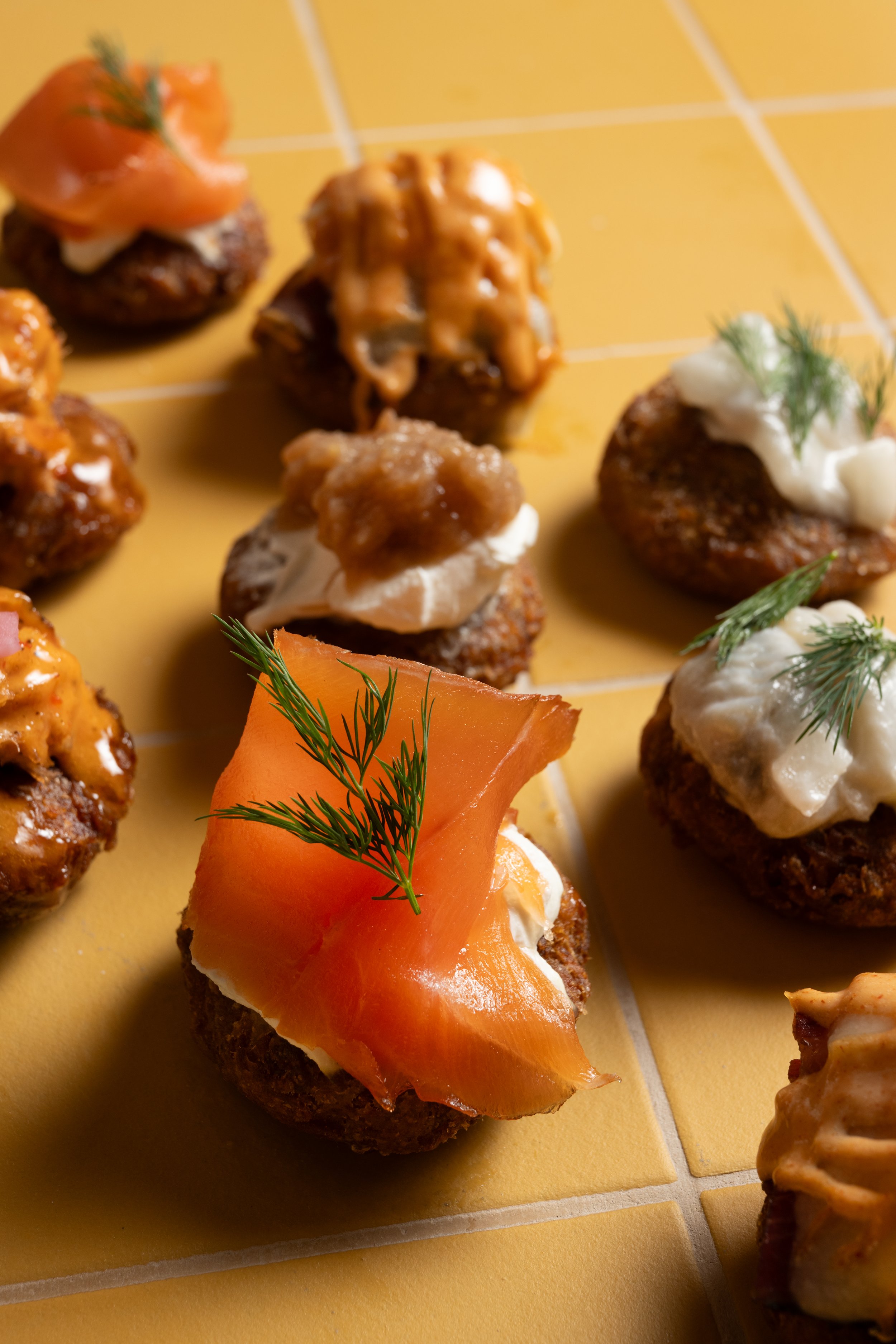 Assorted GERTIE latkes with various toppings including smoked salmon, dill, and creamy sauces on a yellow tiled surface