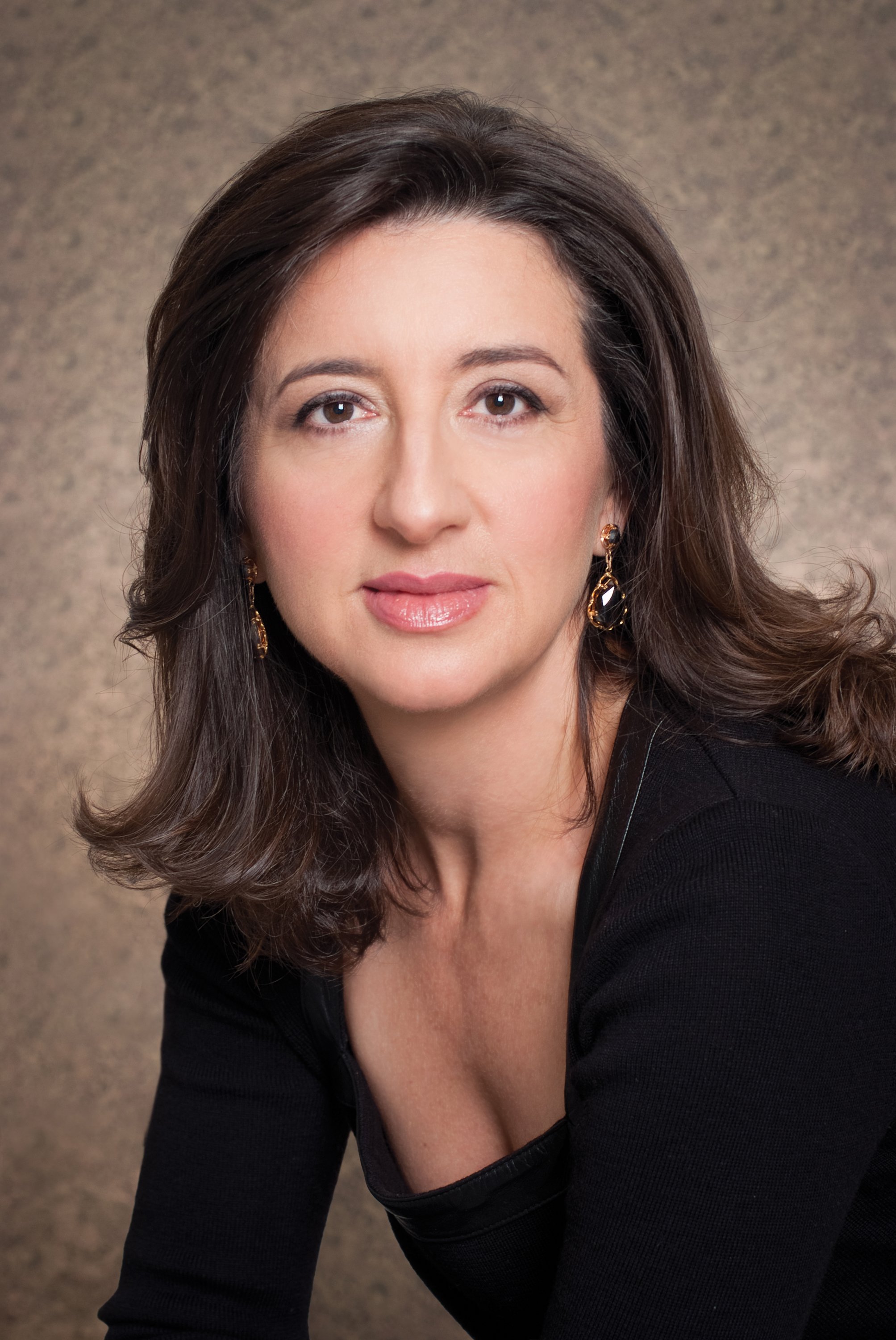 A woman with shoulder-length brown hair, wearing earrings, a black top, and makeup, poses against a textured background.