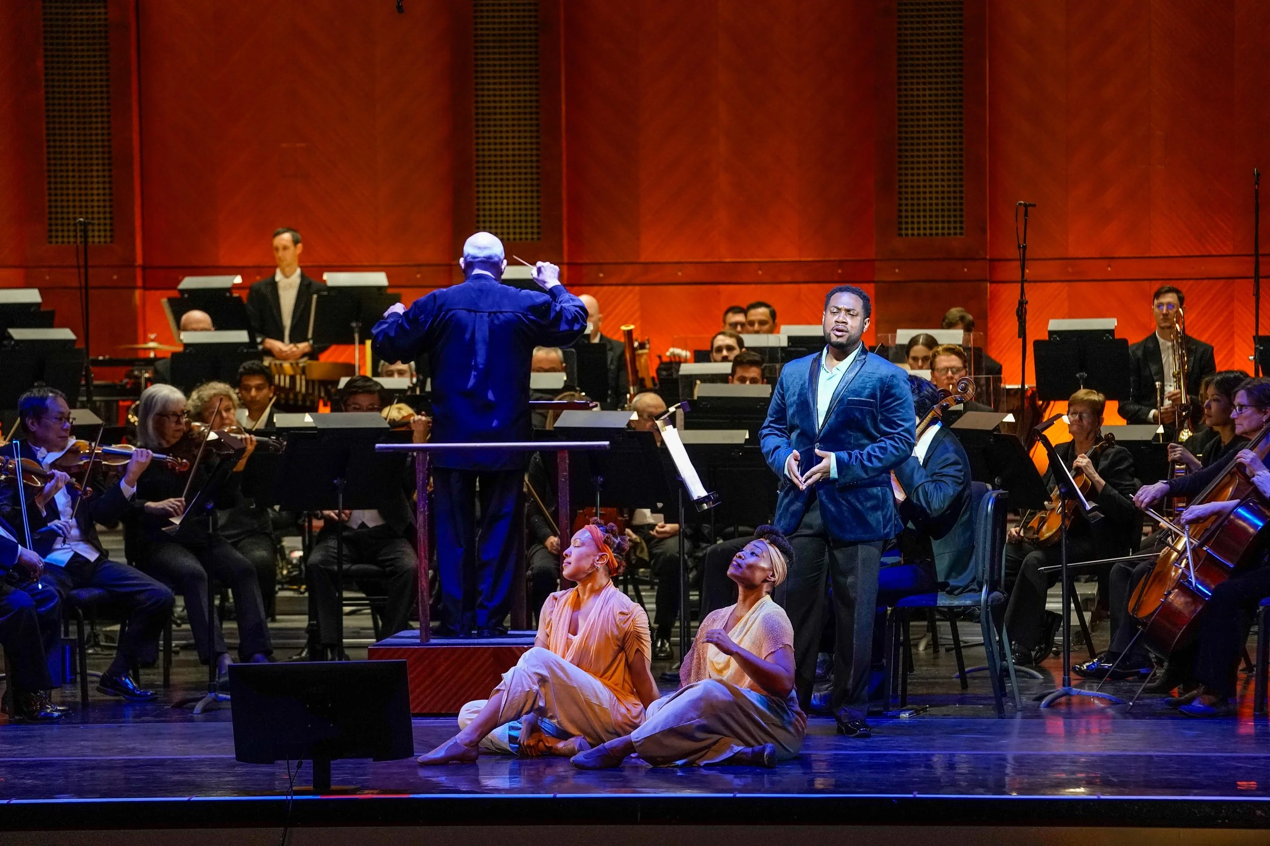 A musical performance on stage with a conductor, orchestra, and performers. Two performers sit on the stage floor in the foreground, one in a beige outfit and the other in a light yellow outfit. A man in a blue velvet jacket is standing and singing, with the orchestra behind them.