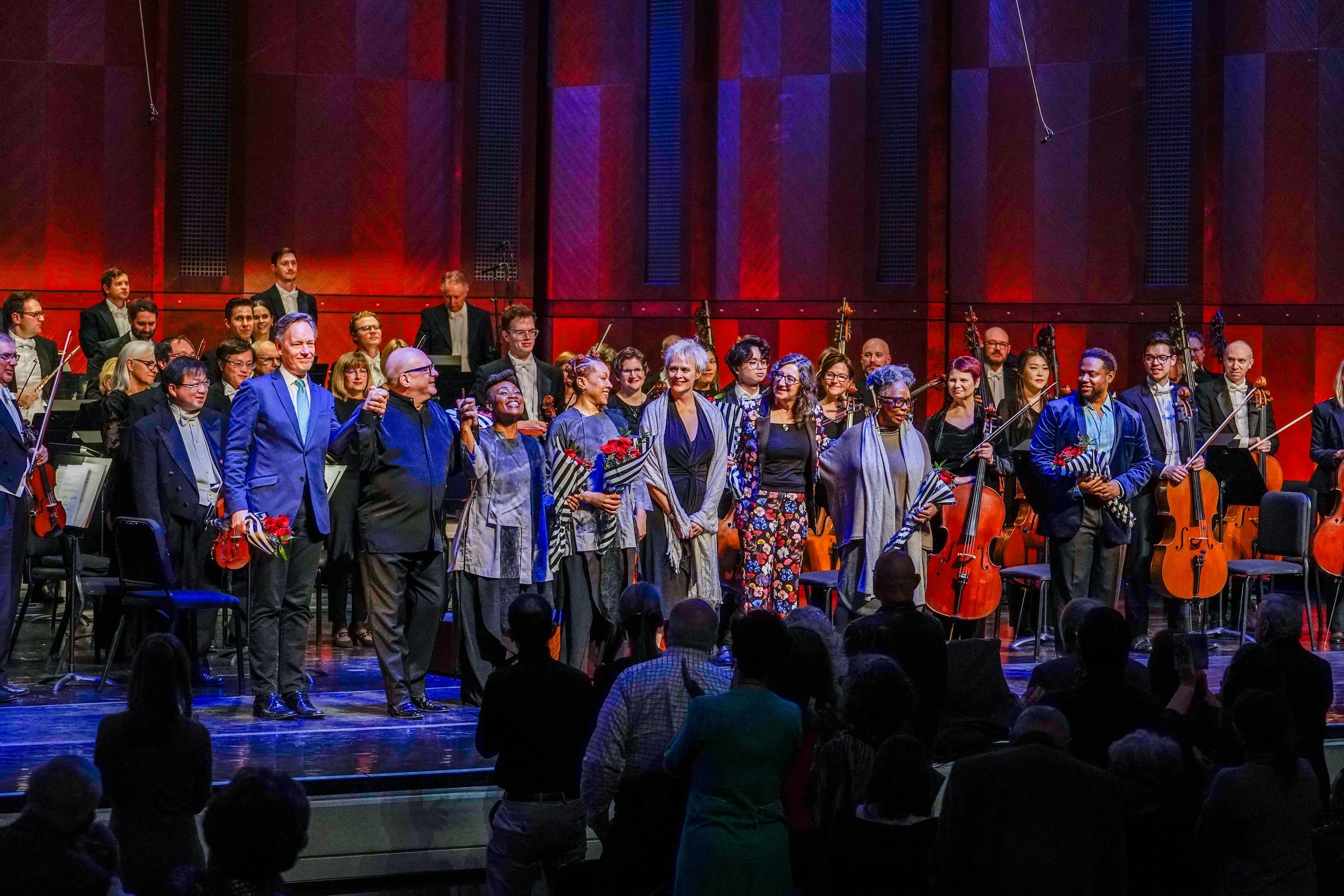 Orchestra on stage after performance, with performers and audience members standing and clapping, some holding flowers.