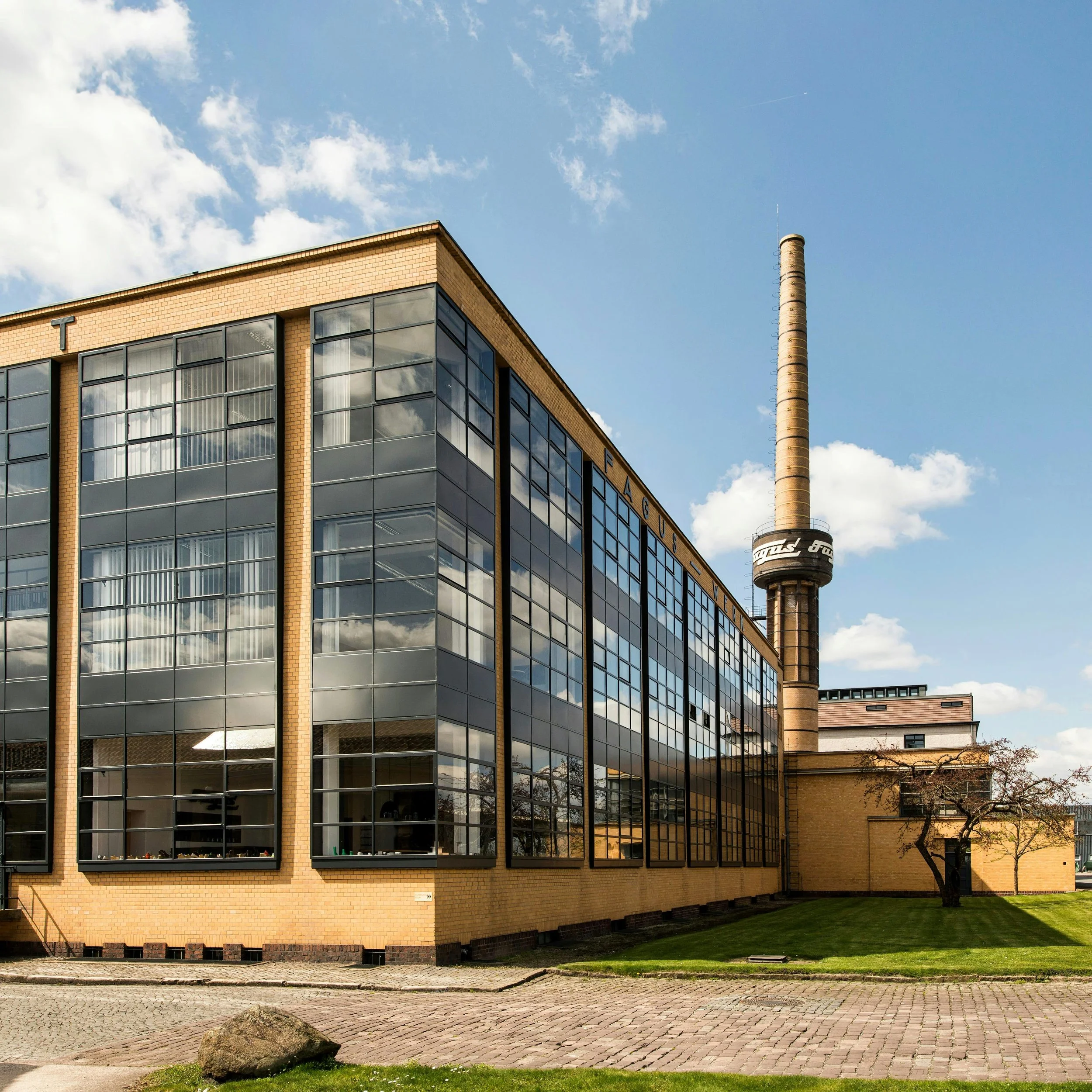 A modern building with large glass windows and beige brick exterior, situated next to a cobblestone street and small grassy area with a large rock in the foreground. A tall smokestack is visible in the background against a partly cloudy blue sky.