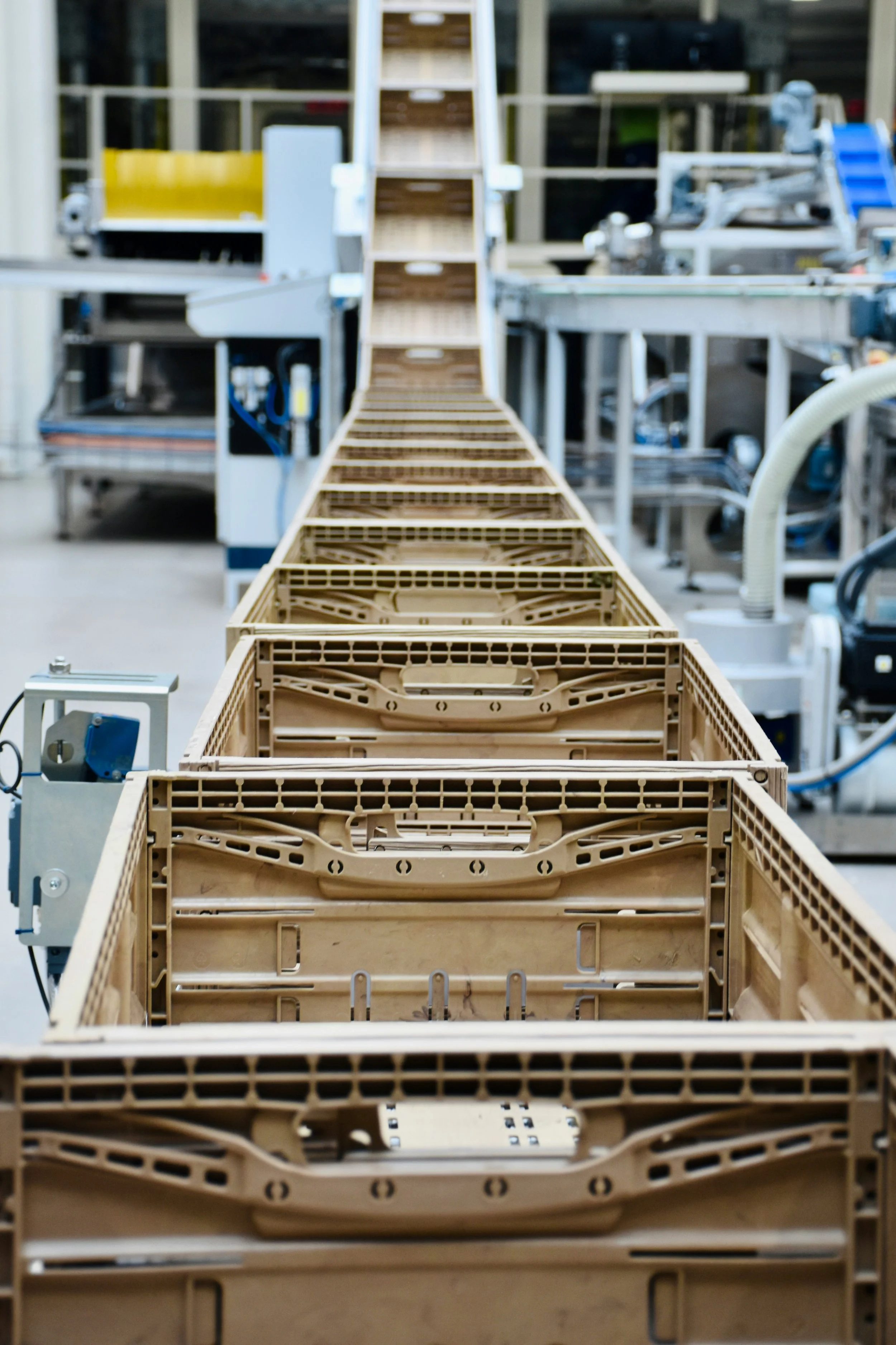 A manufacturing assembly line with empty beige plastic crates moving along a conveyor belt in a factory setting.