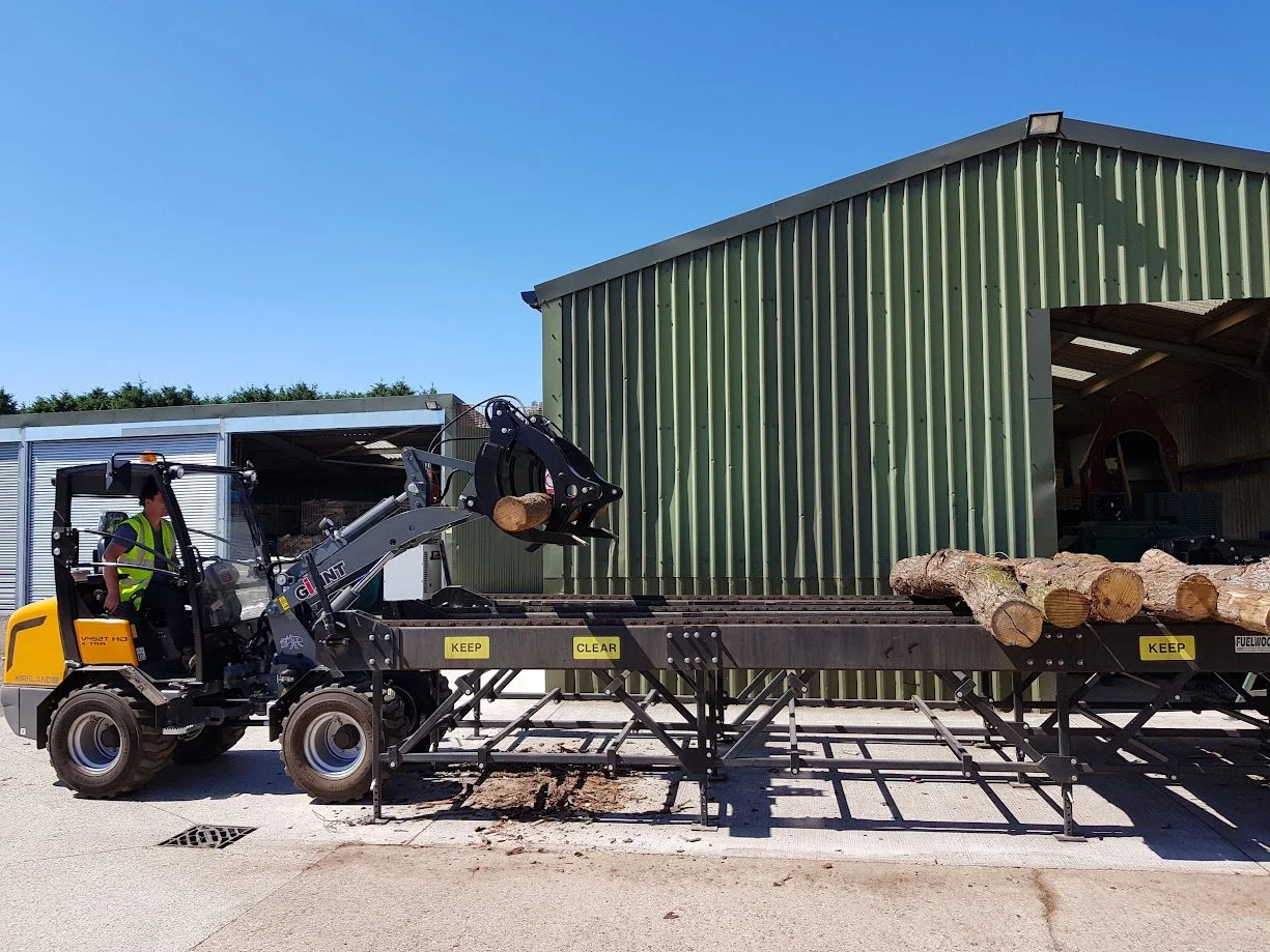 A worker operating a small loader lifting logs onto a flatbed truck nearby a green metal shed under a clear blue sky.