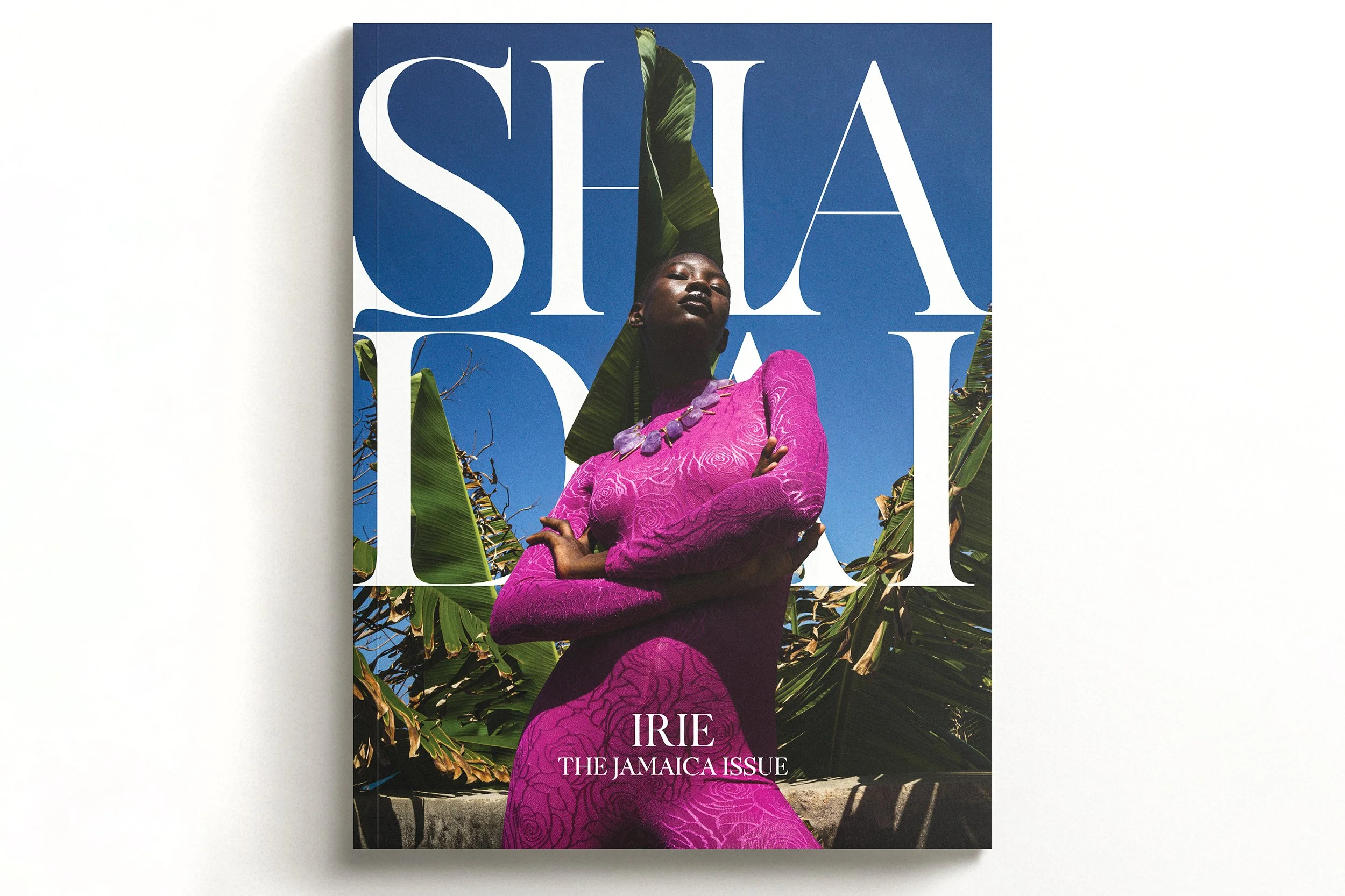 Magazine cover featuring a woman in a vibrant pink dress, standing outdoors among lush green tropical plants, with large white text and a blue sky in the background, titled 'IRE THE JAMAICA ISSUE'.