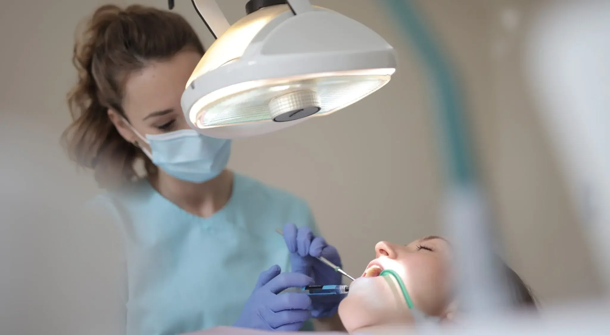 A dental professional in scrubs, mask, and gloves administering anesthesia or sedation to a young patient lying back in a dental chair under a dental light.