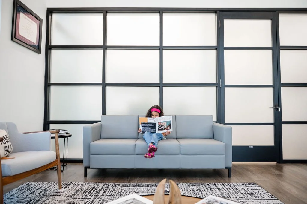 A girl wearing a pink headband and pink shoes sits on a light gray sofa, reading a magazine, in a modern, clean waiting area of a Jacksonville FL dentist office with a black and white patterned rug and glass-paneled wall.