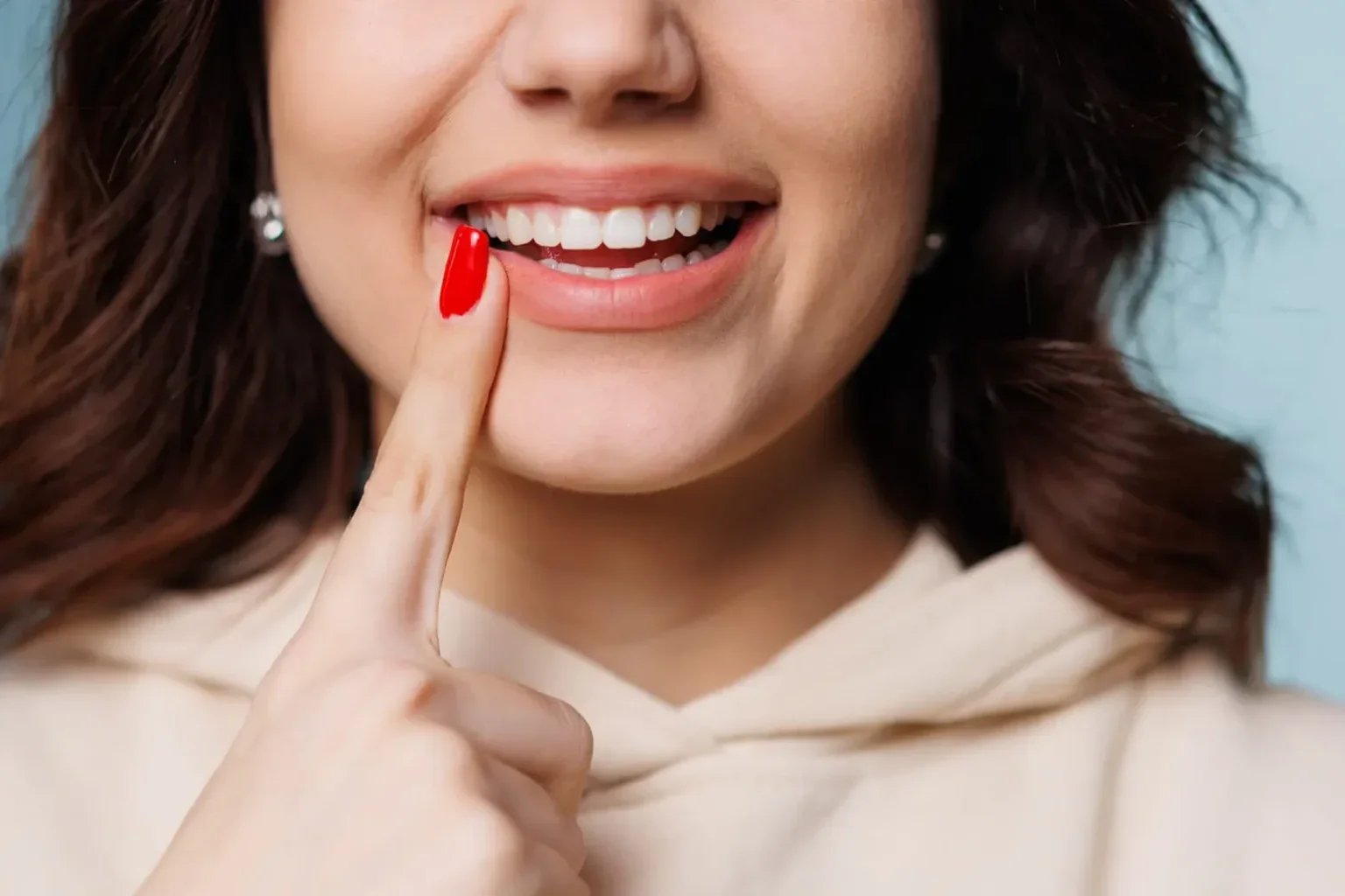 Close-up of a woman smiling and showing straight teeth with her finger on her cheek, wearing red nail polish and earrings.