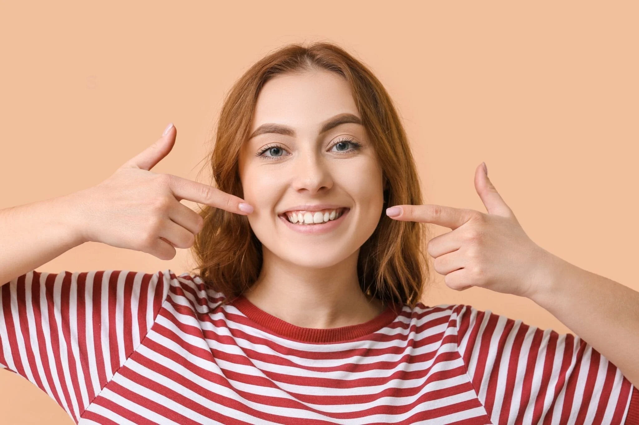 A young woman with red hair and blue eyes smiling, pointing to her cheeks with both index fingers, wearing a red and white striped shirt, standing against a peach-colored background.