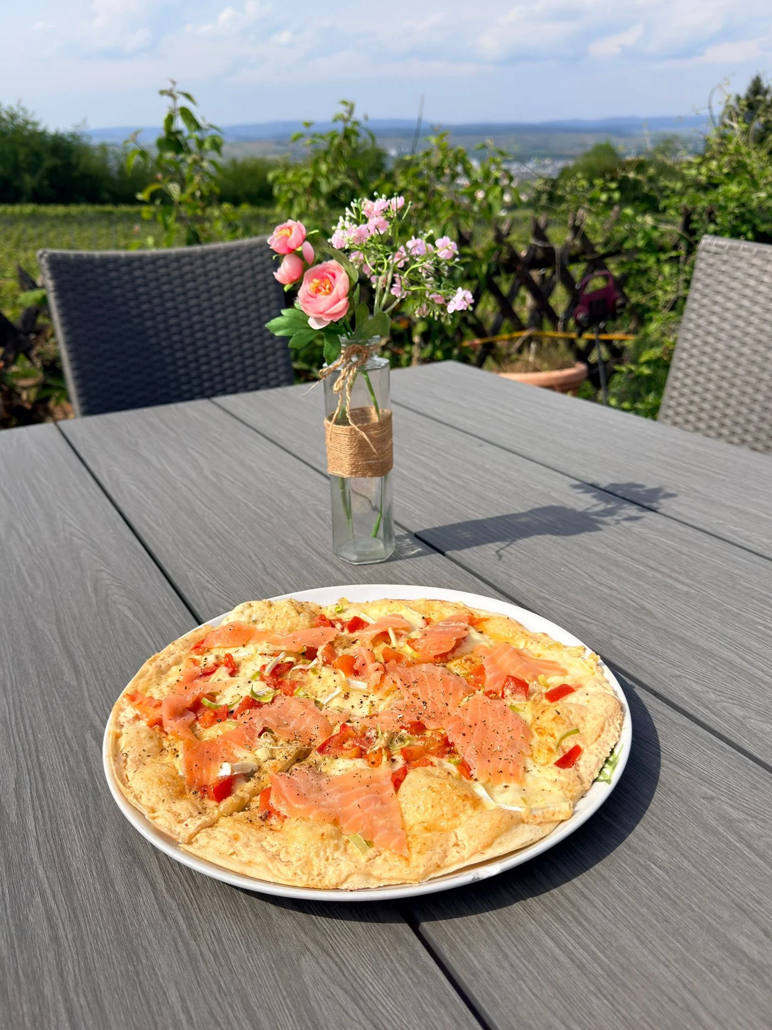 Pizzastück mit Lachs, Tomaten und Käse auf weißem Teller auf einem grauen Tisch im Freien, im Hintergrund grüne Büsche, Wiesen und ein blauer Himmel mit Wolken.