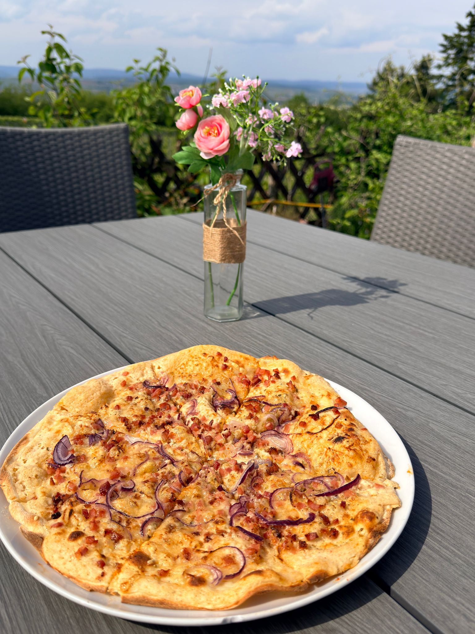 Mittagsessen mit einer Pizza auf einem weißen Teller im Freien, im Hintergrund ein Tisch mit einer Vase mit pinken Blumen auf einer Terrasse mit grünen Büschen und einem Blick auf den Himmel.